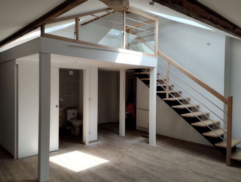 Chambre mansardée avec des poutres apparentes, un escalier et un petit coin salle de bains dans un intérieur partiellement aménagé