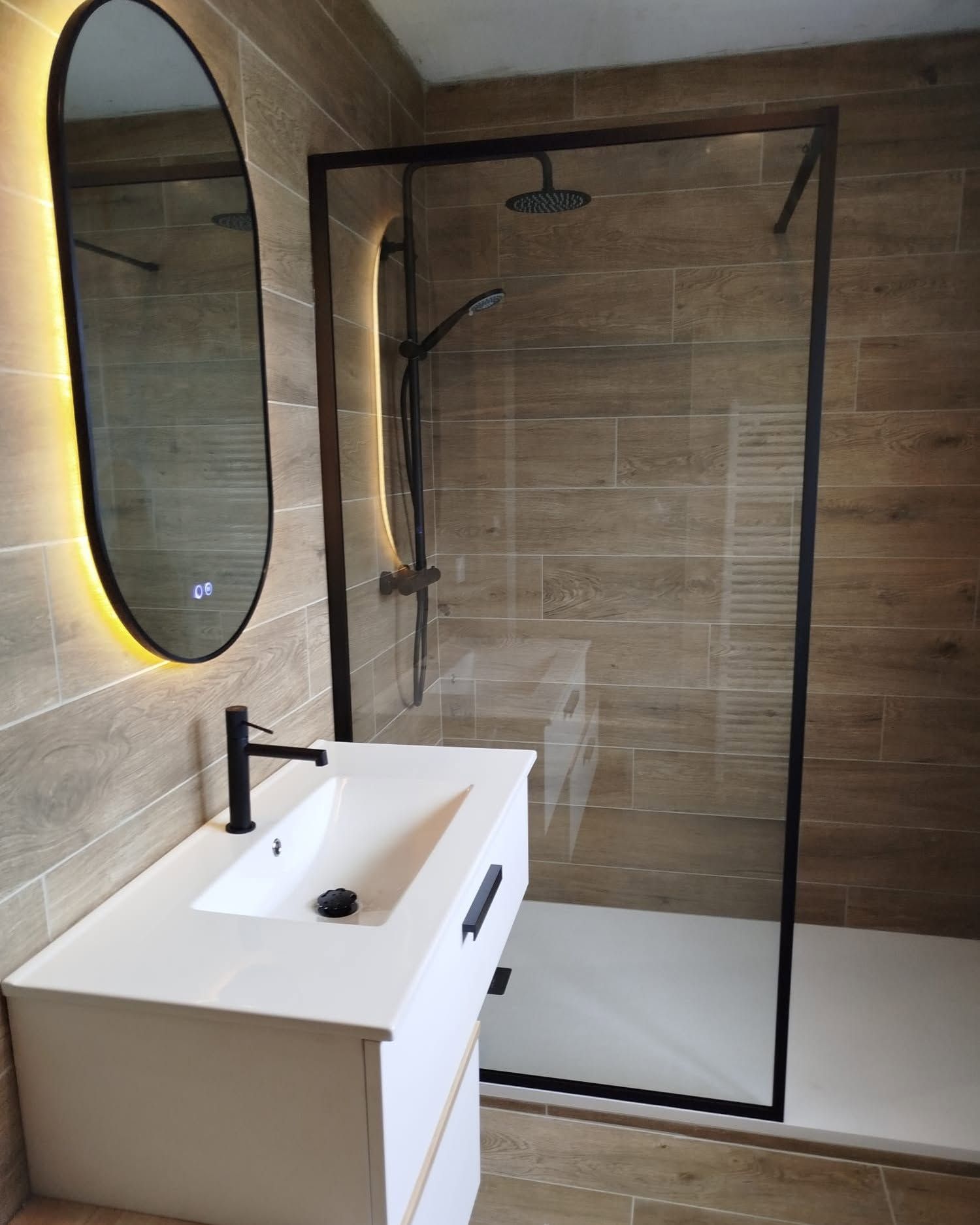 Salle de bains moderne avec une douche en verre, un lavabo blanc, un miroir ovale et des murs carrelés en imitation bois