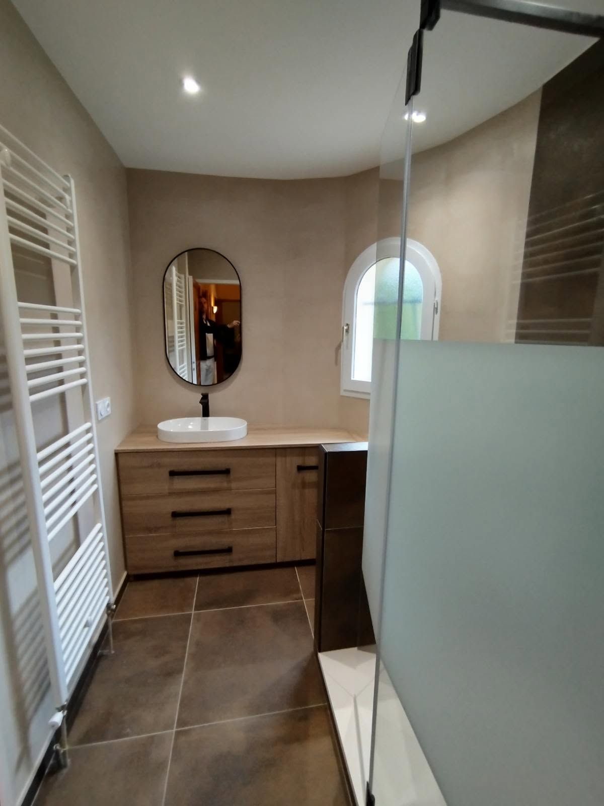 Salle de bains moderne avec un meuble de lavabo en bois, un miroir ovale, carrelage marron et cabine de douche en verre dépoli