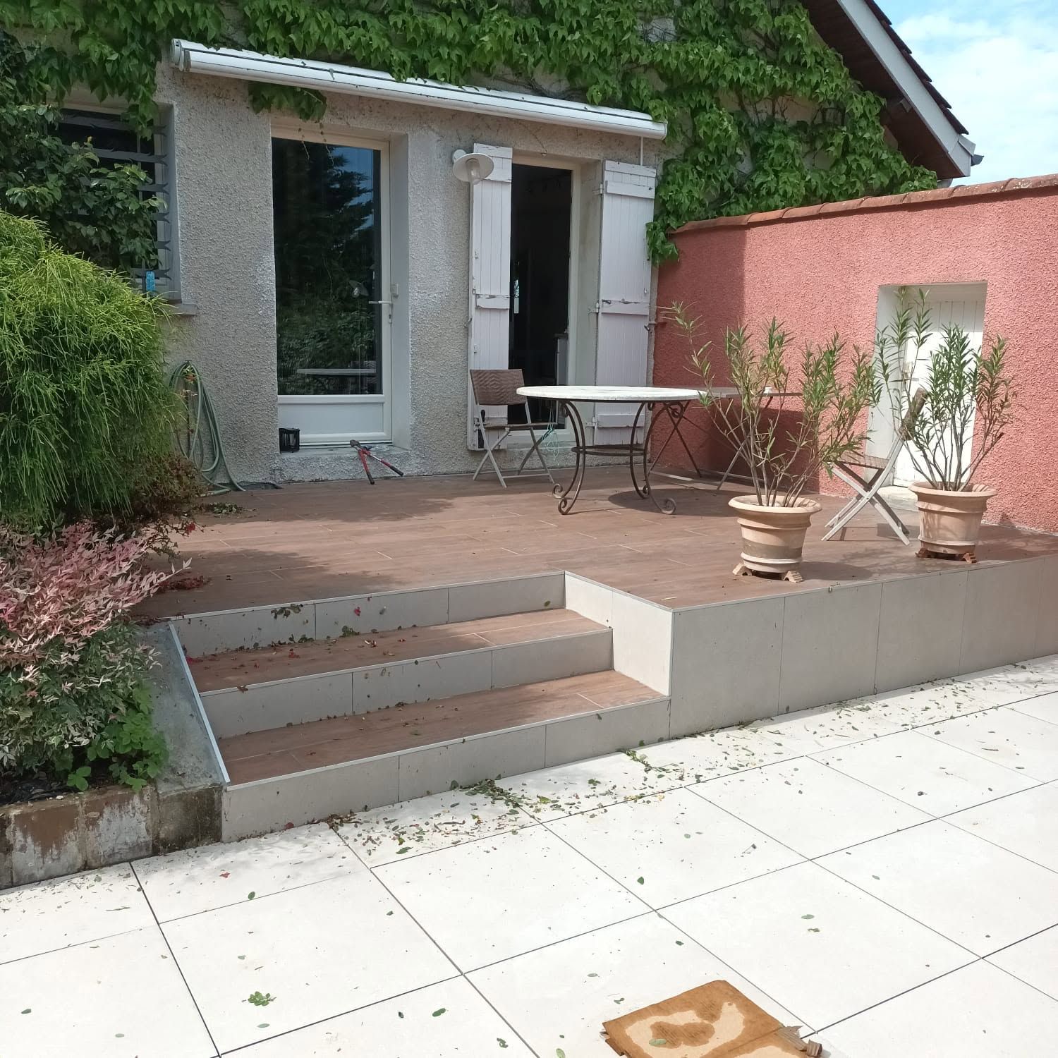 Petit patio avec marches, dallage blanc, plantes en pot et une table à l'extérieur d'une porte, à côté d'un mur couleur pêche.