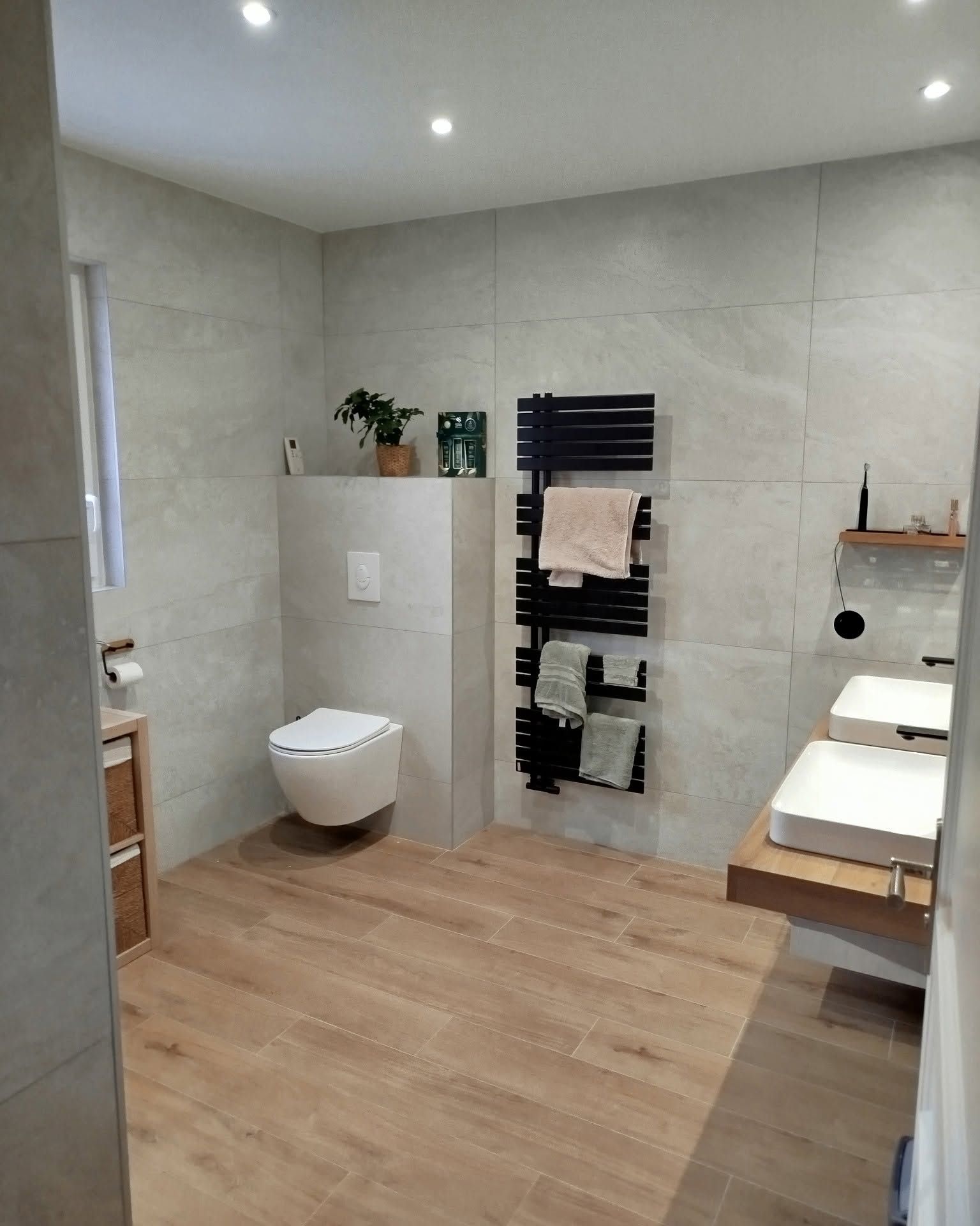 Salle de bains moderne avec un sol en imitation bois, des toilettes suspendues, un porte-serviettes noir et des lavabos blancs.