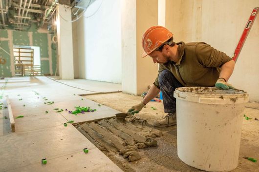 Un ouvrier du bâtiment portant un casque orange étale du béton frais sur le sol à l'intérieur d'un bâtiment.
