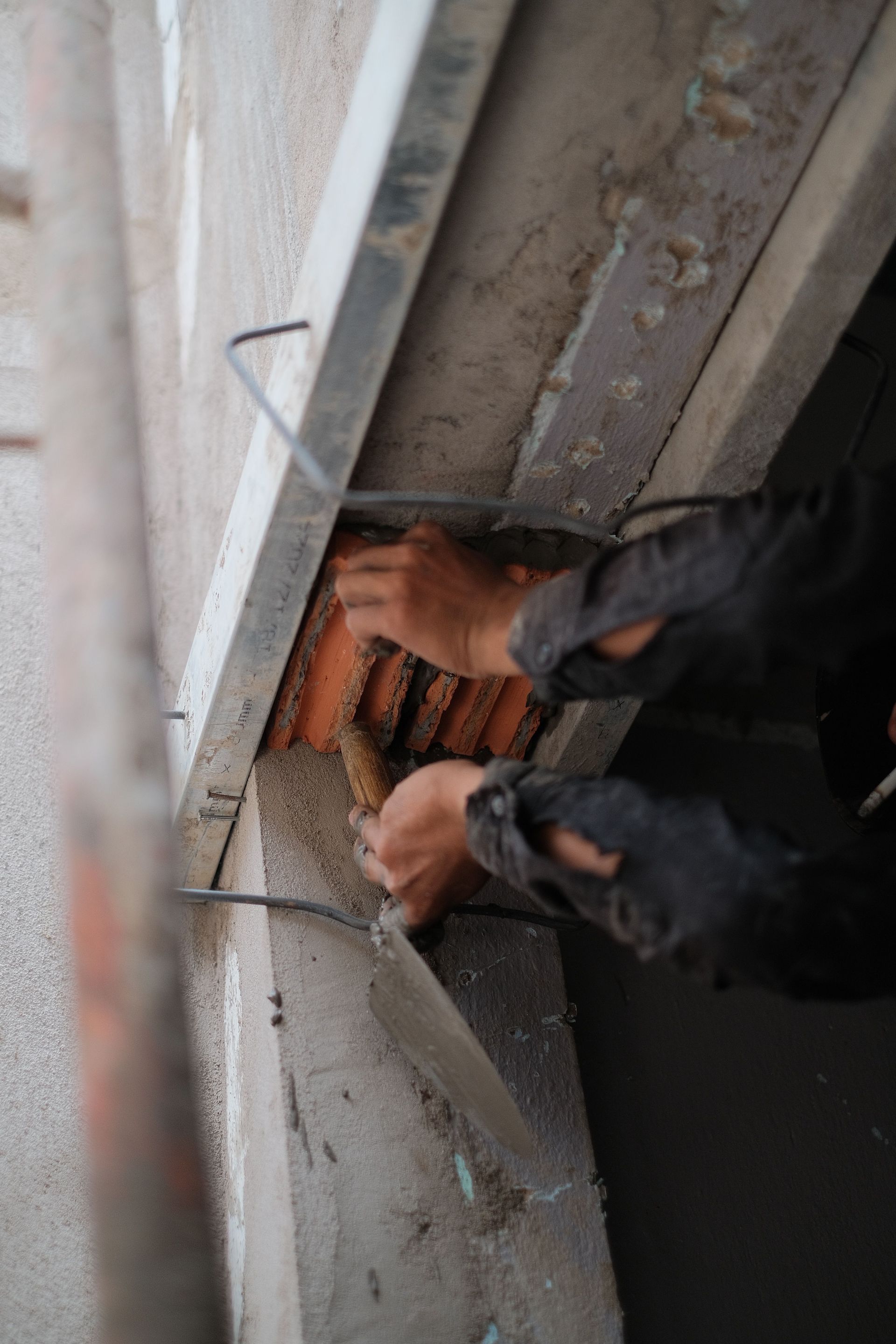 Des mains installent des briques rouges dans une petite ouverture d'un mur en béton