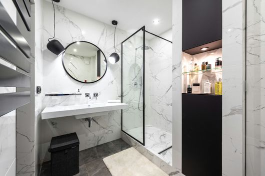 Salle de bains moderne avec des murs en marbre, un miroir rond, un lavabo blanc, une douche à cadre noir et une poubelle noire