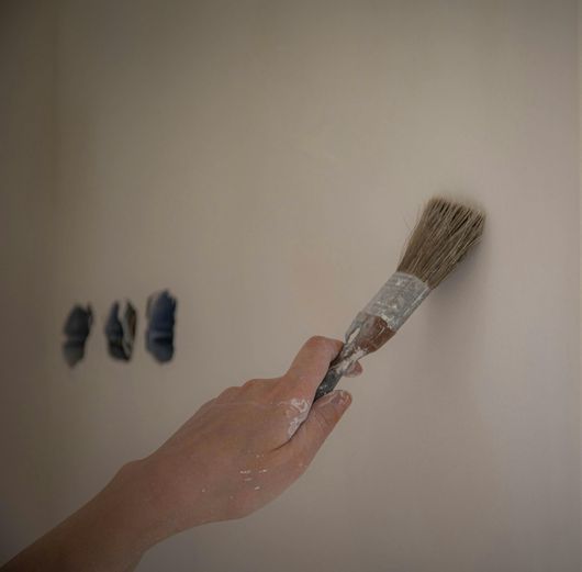 Une main tient un pinceau en train de peindre un mur beige, avec trois prises murales bleues sur la gauche.