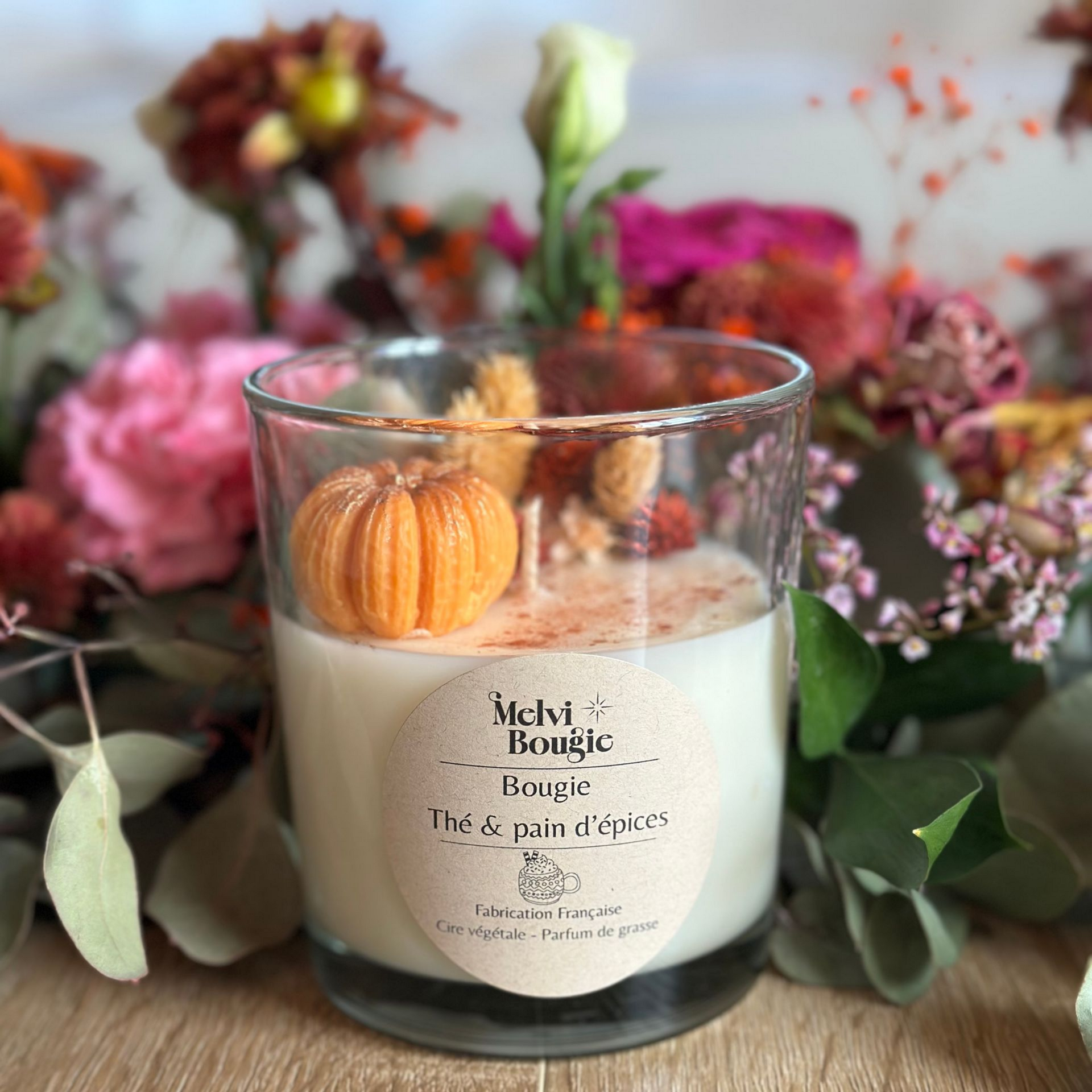 Bougie dans un verre, décorée d'une citrouille, avec des fleurs en arrière-plan.