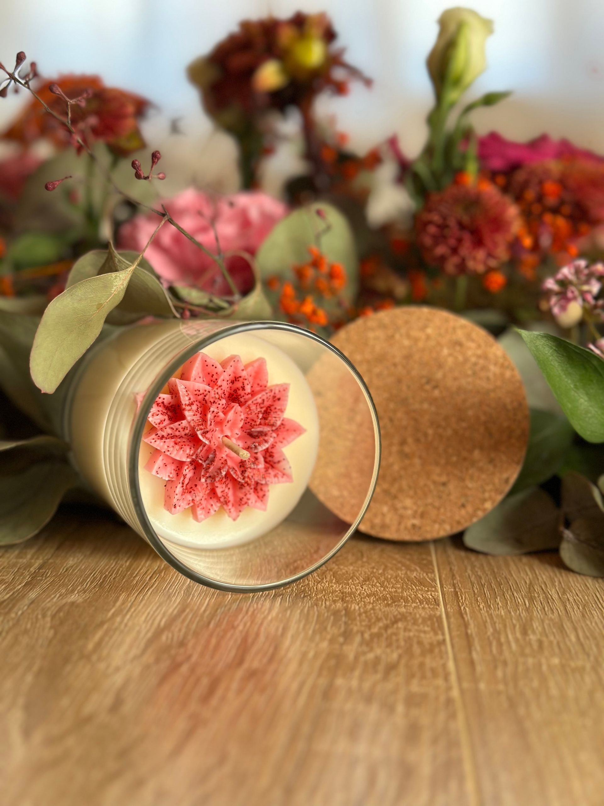 Bougie dans un verre, ornée d'un motif floral rose, posée sur une surface en bois, avec des fleurs en arrière-plan.