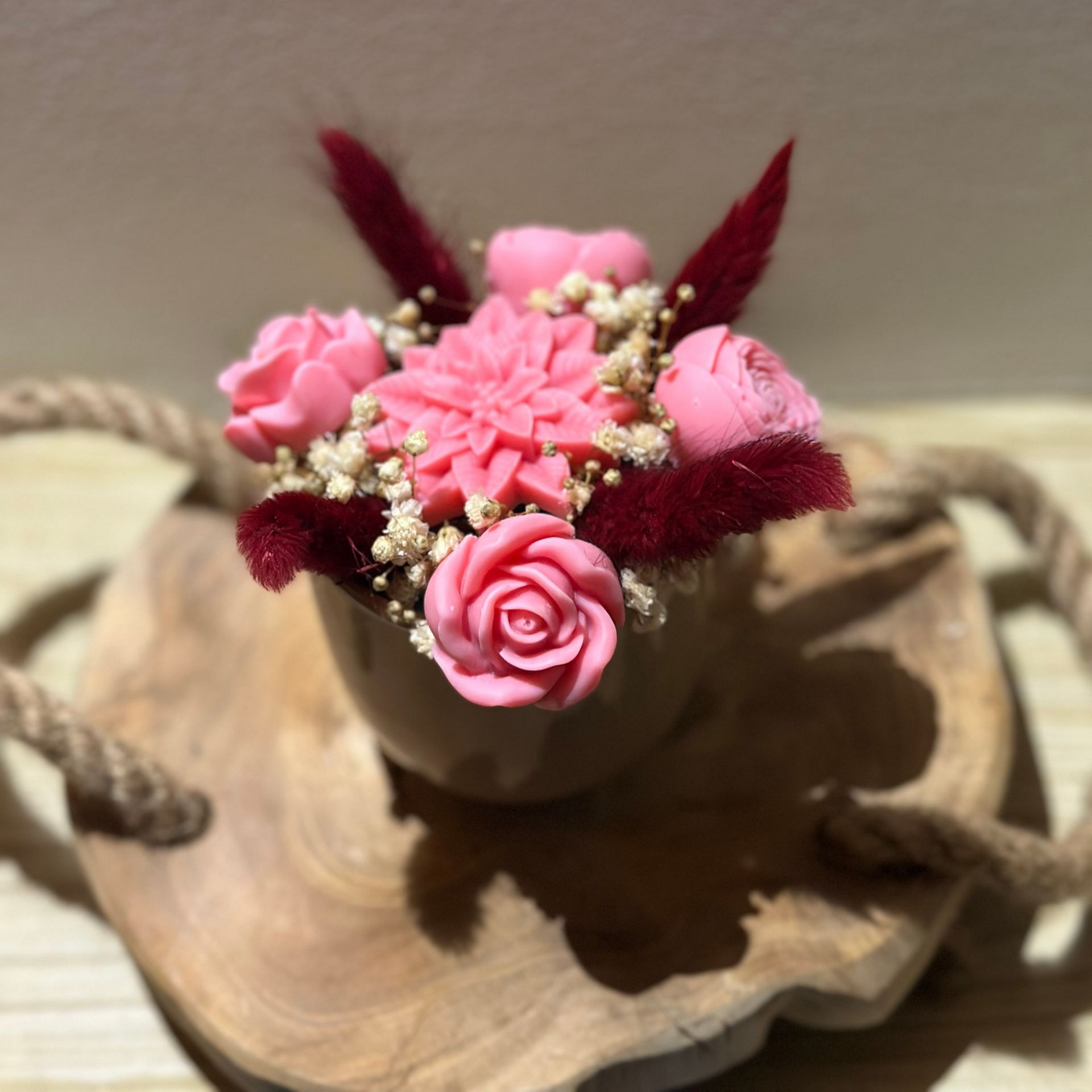 Bouquet de fleurs de cire rose dans un récipient en bois, posé sur un plateau en bois avec des poignées en corde.