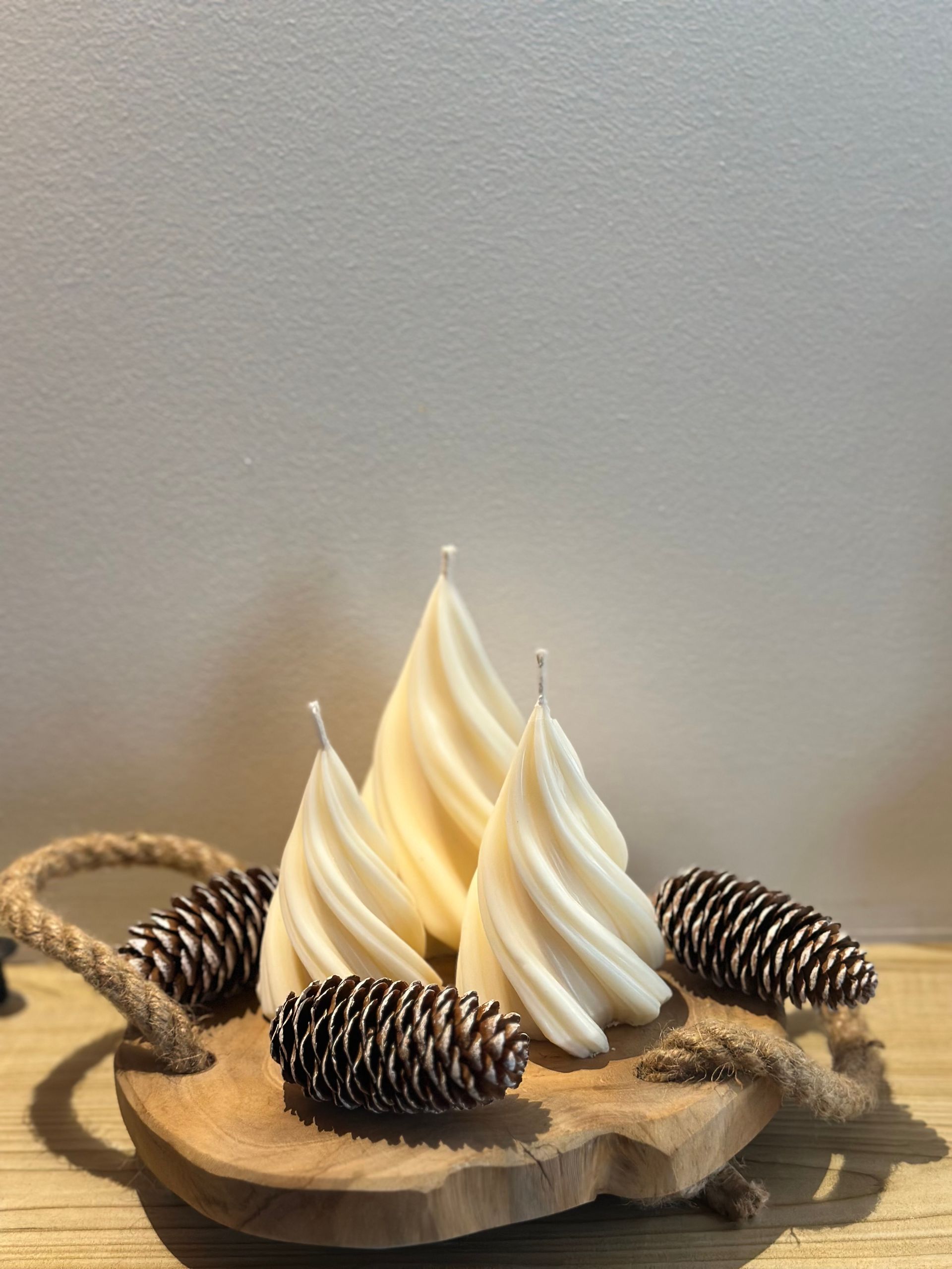 Trois bougies crème disposées en spirale et des pommes de pin sur un plateau en bois, détail de corde, sur un mur blanc.