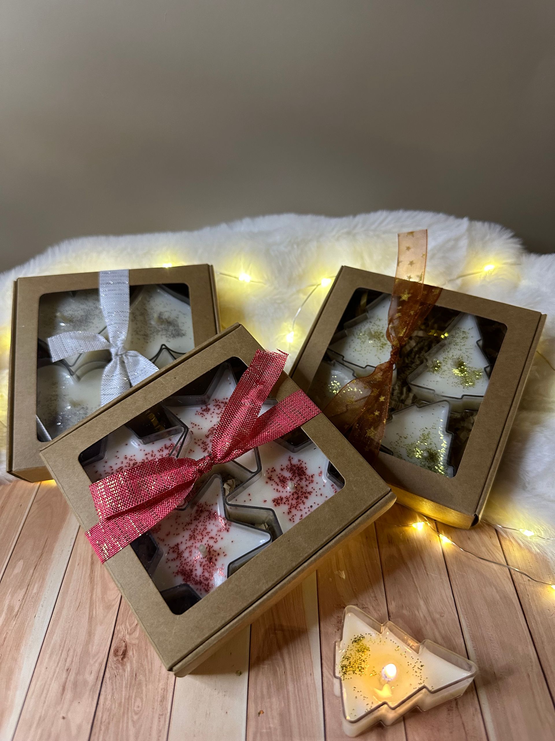 Trois boîtes de fondants parfumés en forme d'étoile, attachées par un ruban, sont posées sur une surface avec des guirlandes lumineuses et un seul fondant parfumé allumé.