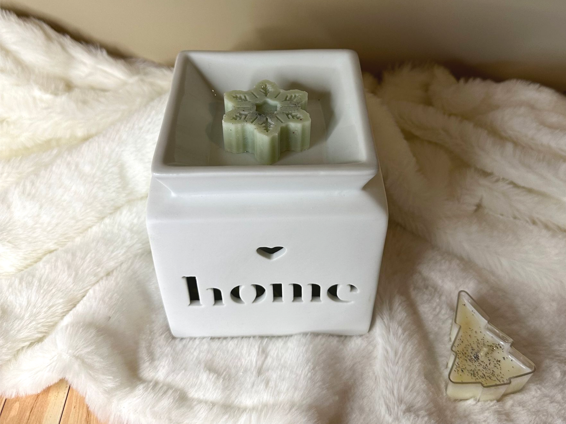 Chauffe-cire blanc «maison» avec fondants parfumés en forme de flocons de neige et de sapins de Noël sur une couverture moelleuse.