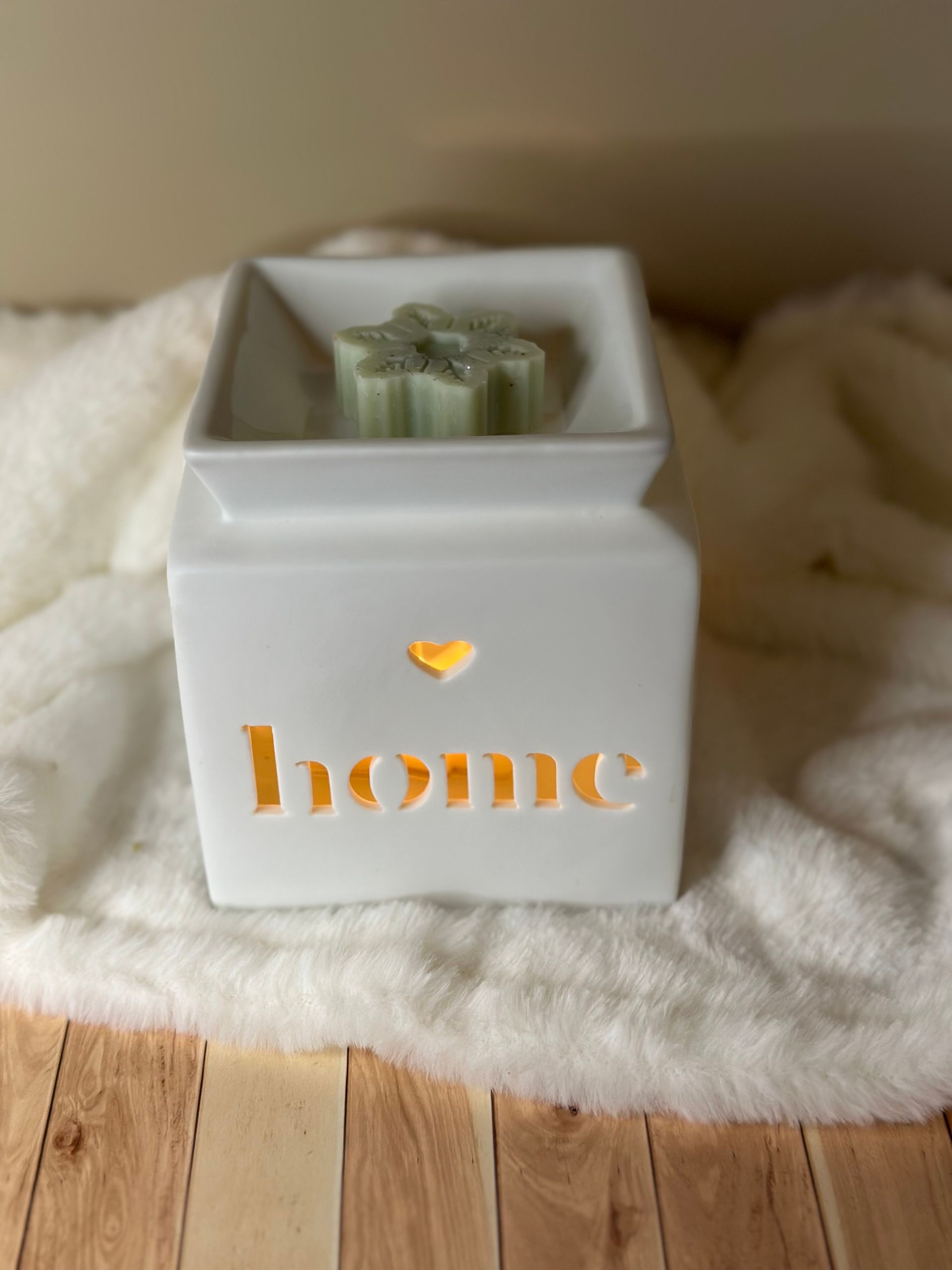 Chauffe-cire en céramique blanche avec découpe « maison », illuminé, et contient une cire verte fondante. Posé sur un tapis blanc moelleux.