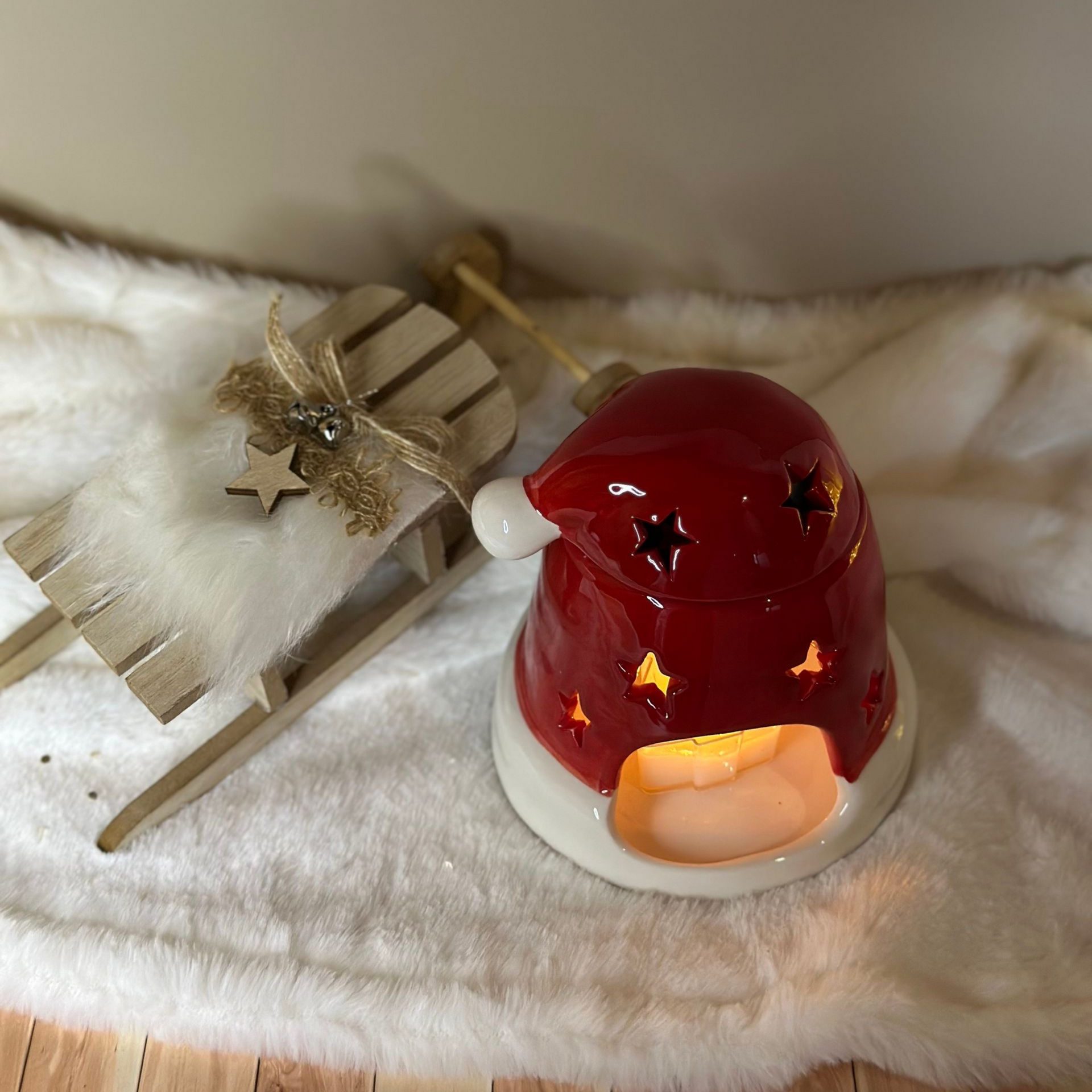 Un bougeoir en céramique en forme de bonnet de père Noël, orné d'étoiles découpées, est placé à côté d'un petit traîneau en bois sur une surface blanche et moelleuse.