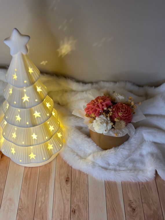 Sapin de Noël en céramique illuminé à côté d'une composition florale sur une surface blanche et moelleuse.