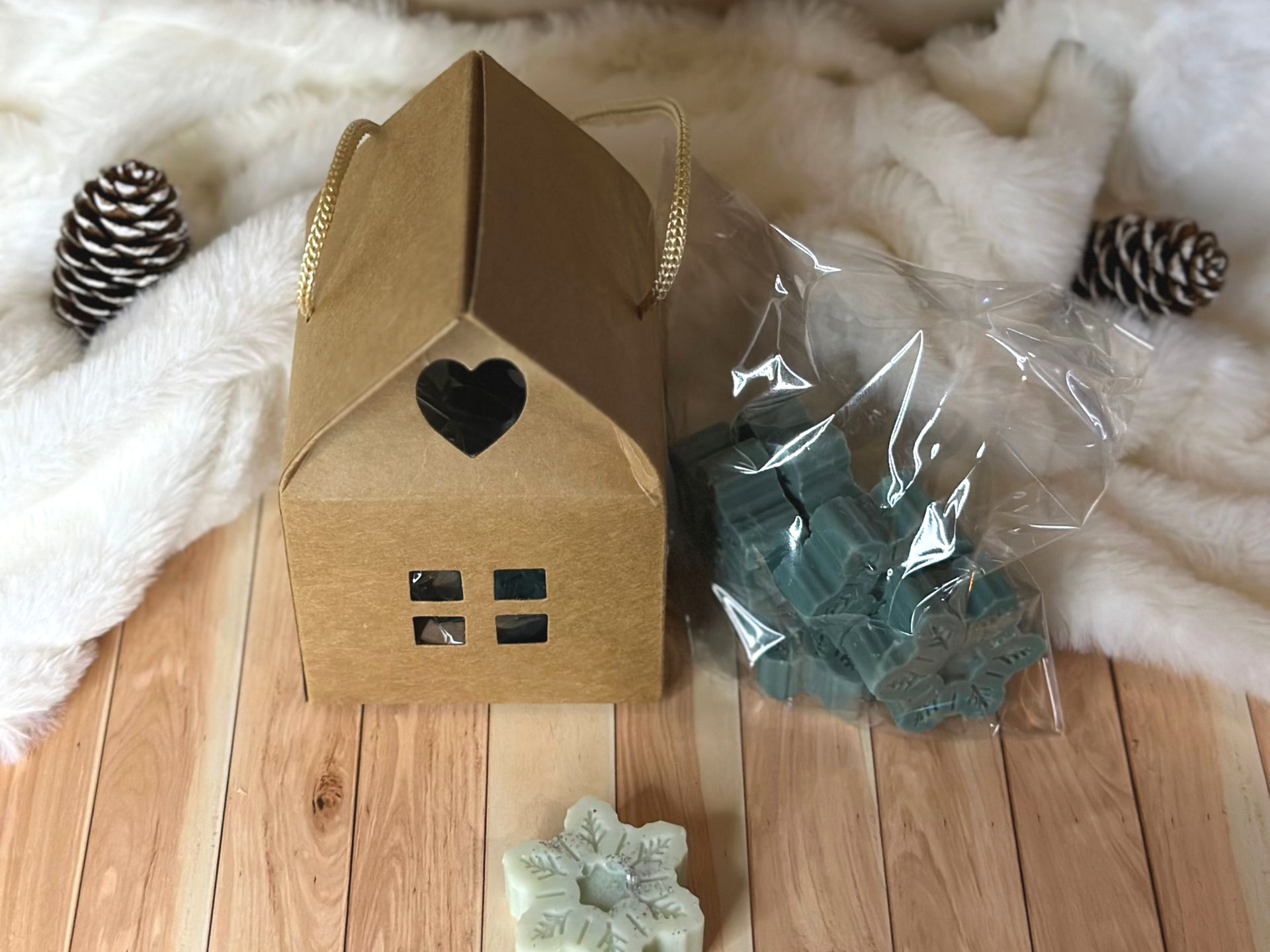 Boîte cadeau marron en forme de maison avec une découpe en forme de cœur et un sachet de fondants parfumés turquoise, sur un fond blanc duveteux orné de pommes de pin.