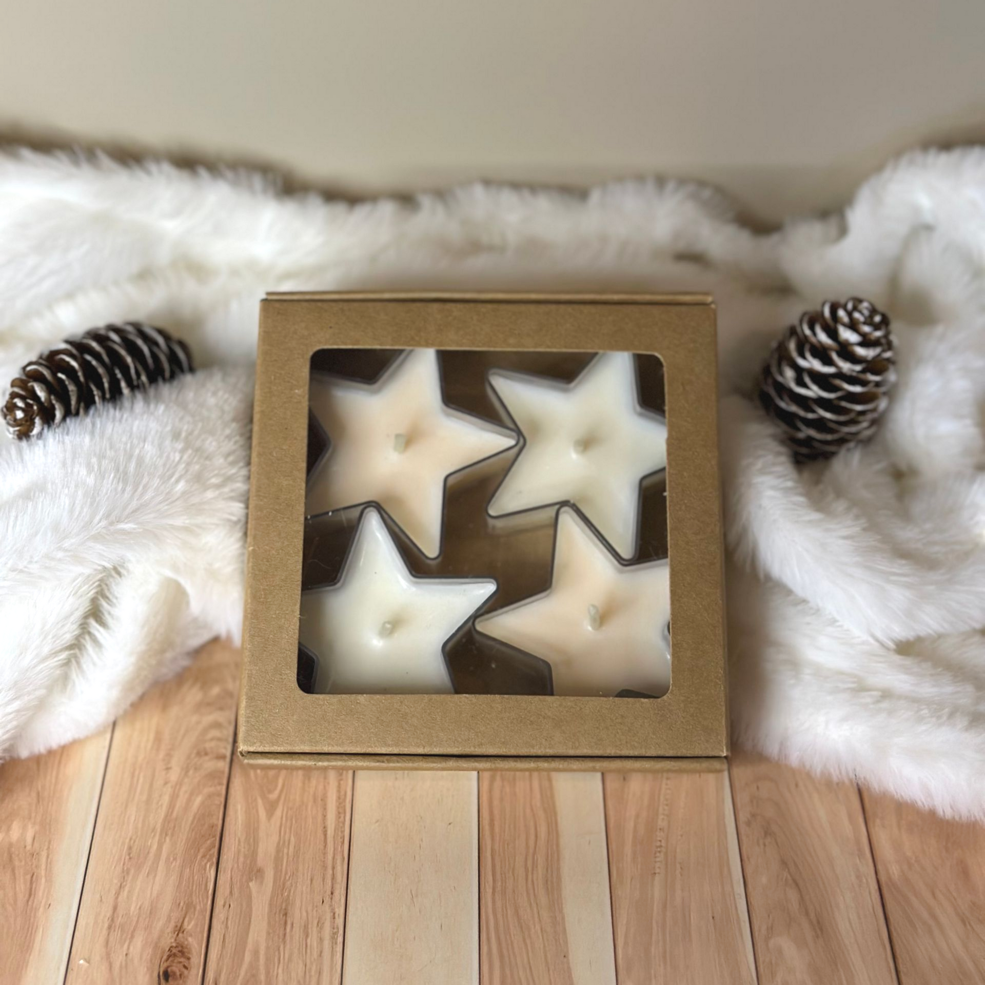 Des bougies en forme d'étoile dans une boîte avec une fenêtre, sur une surface blanche duveteuse, et un fond en bois avec des pommes de pin.