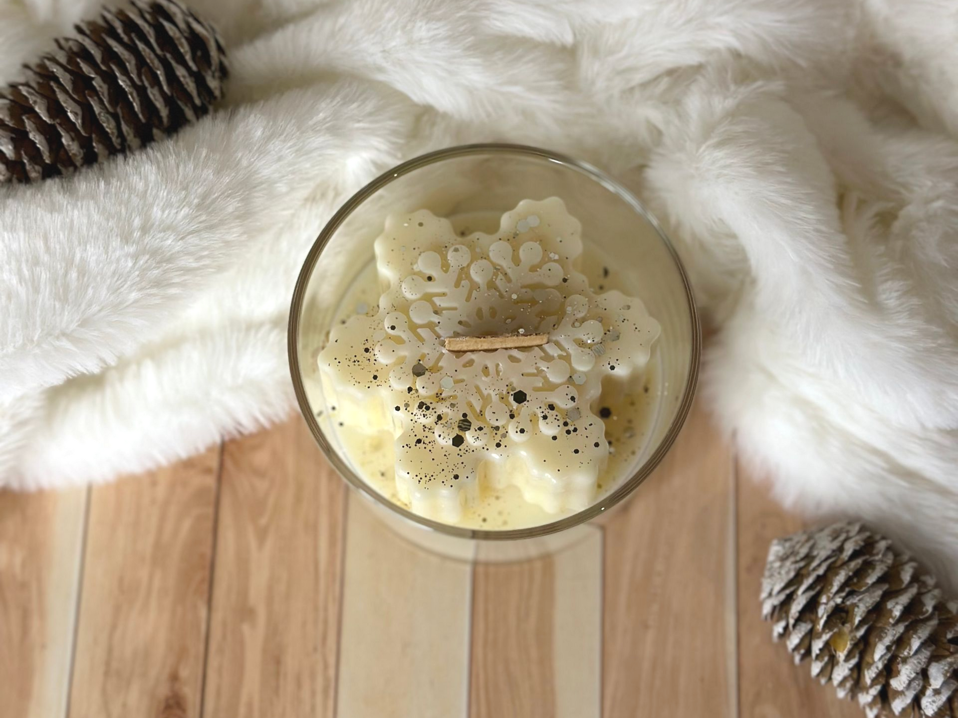 Bougie en forme de flocon de neige dans un bocal en verre, posée sur une fourrure blanche duveteuse ornée de pommes de pin, sur une surface en bois.
