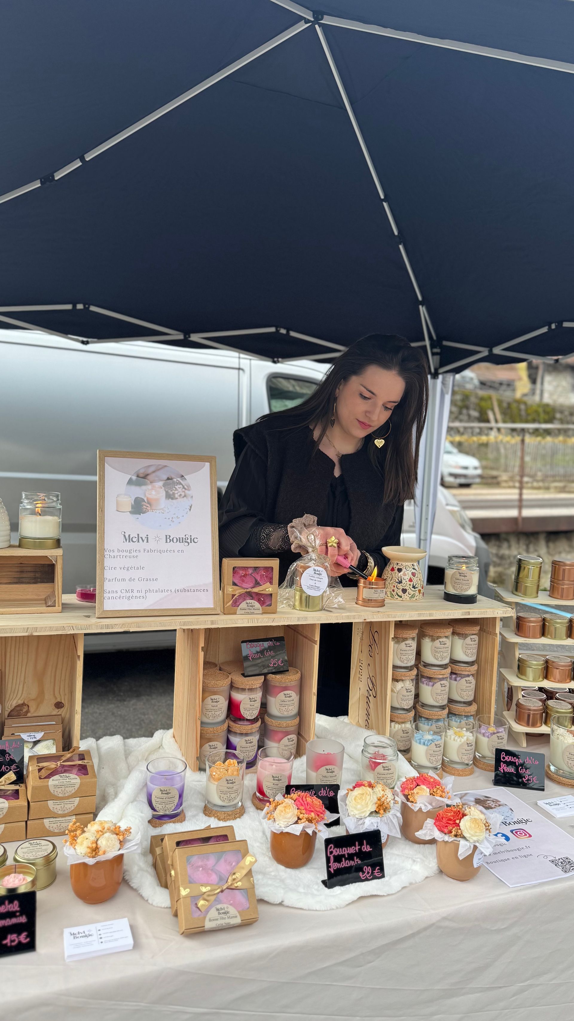 Bougie dans un verre, ornée d'un motif floral rose, posée sur une surface en bois, avec des fleurs en arrière-plan.