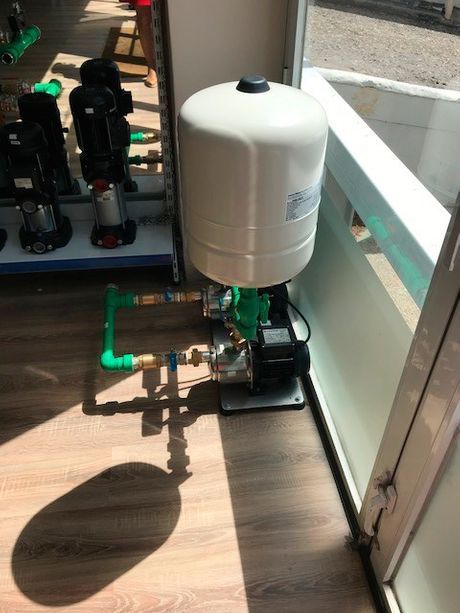 Sistema de bomba de presión de agua, con tanque y tuberías verdes, sobre un piso de madera cerca de una ventana.