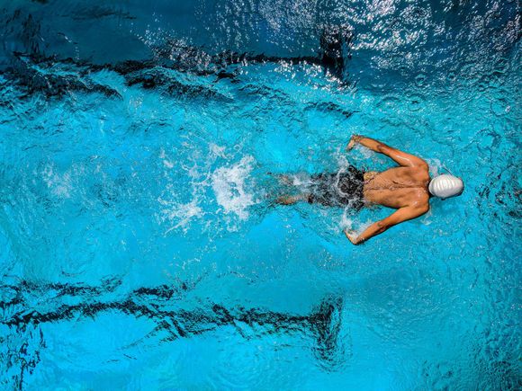 Nadador en una piscina azul, vista aérea. Brazos extendidos, impulsándose en el agua, con gorra blanca y bañador negro.