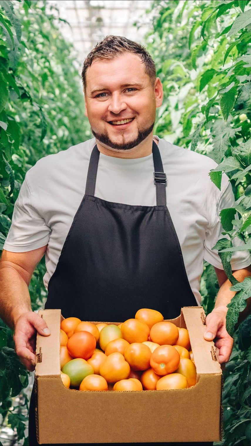 Una persona sonriente con un delantal negro sostiene una caja de cartón llena de tomates frescos mientras está de pie en un invernadero.