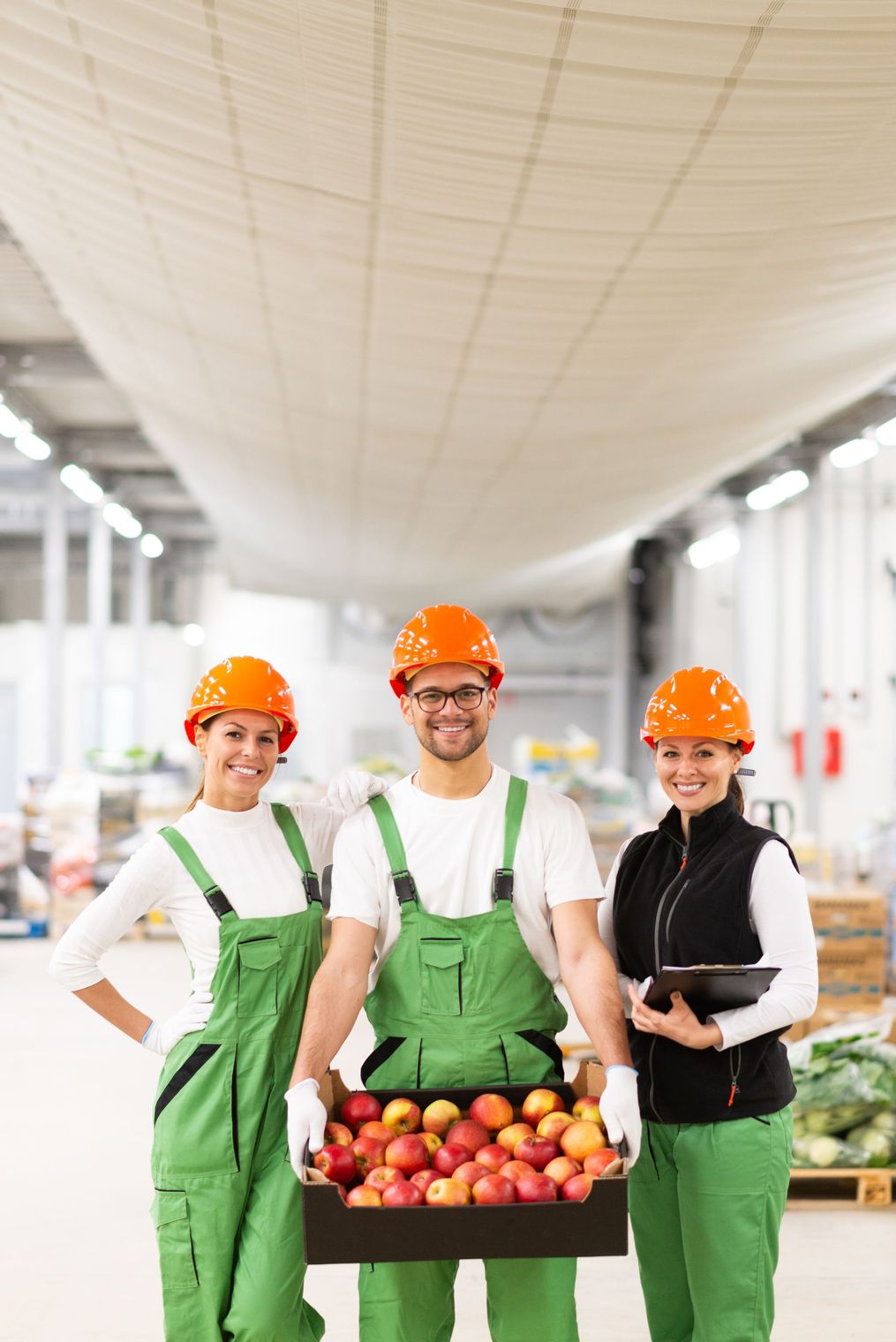 Tres personas con cascos naranjas y ropa de trabajo verde están de pie en un almacén; la persona del centro sostiene una caja de manzanas.