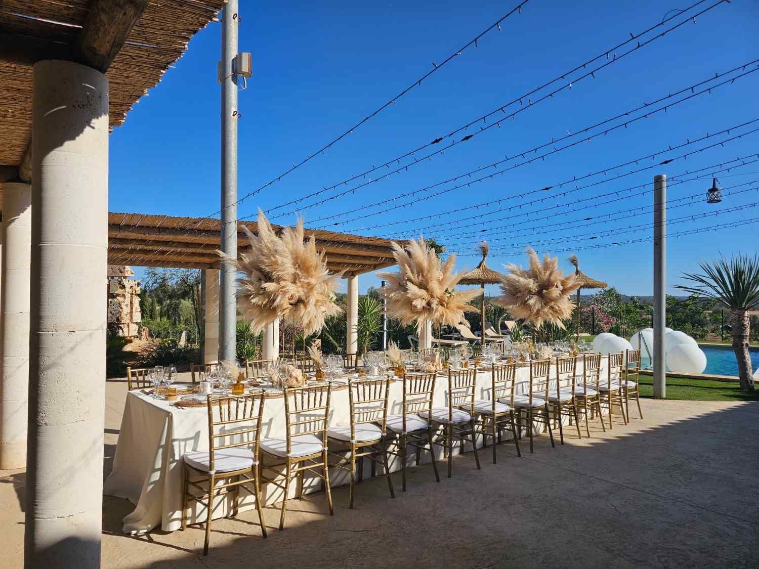 Comedor al aire libre con mesas y sillas blancas bajo una pérgola, junto al mar azul y un cielo despejado.
