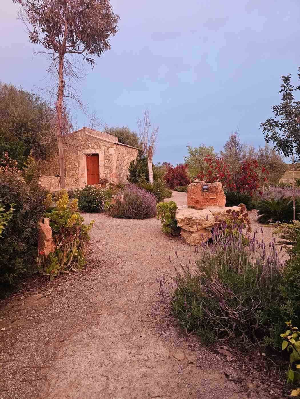 Sendero de piedra que atraviesa un jardín hasta una pequeña cabaña rústica, con arbustos y árboles bajo un cielo rosado.