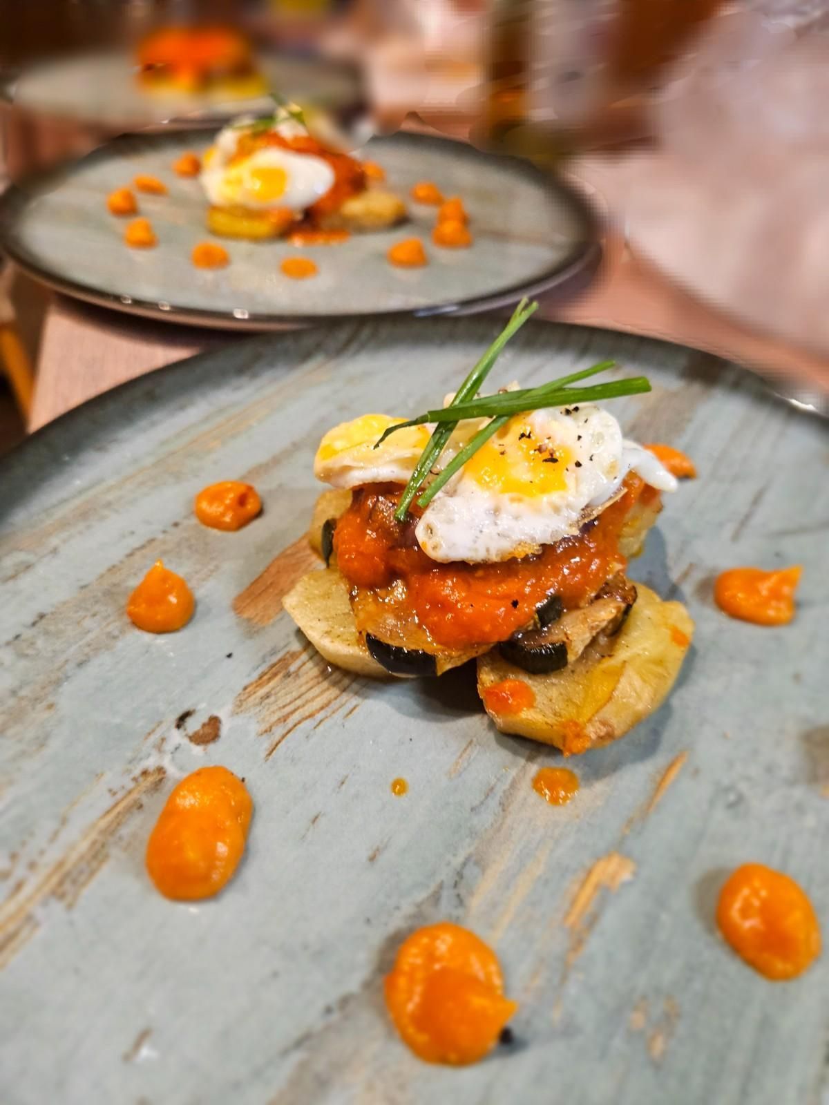 Aperitivo gourmet en un plato gris, coronado con huevo y hierbas, adornado con puntos de salsa de naranja.