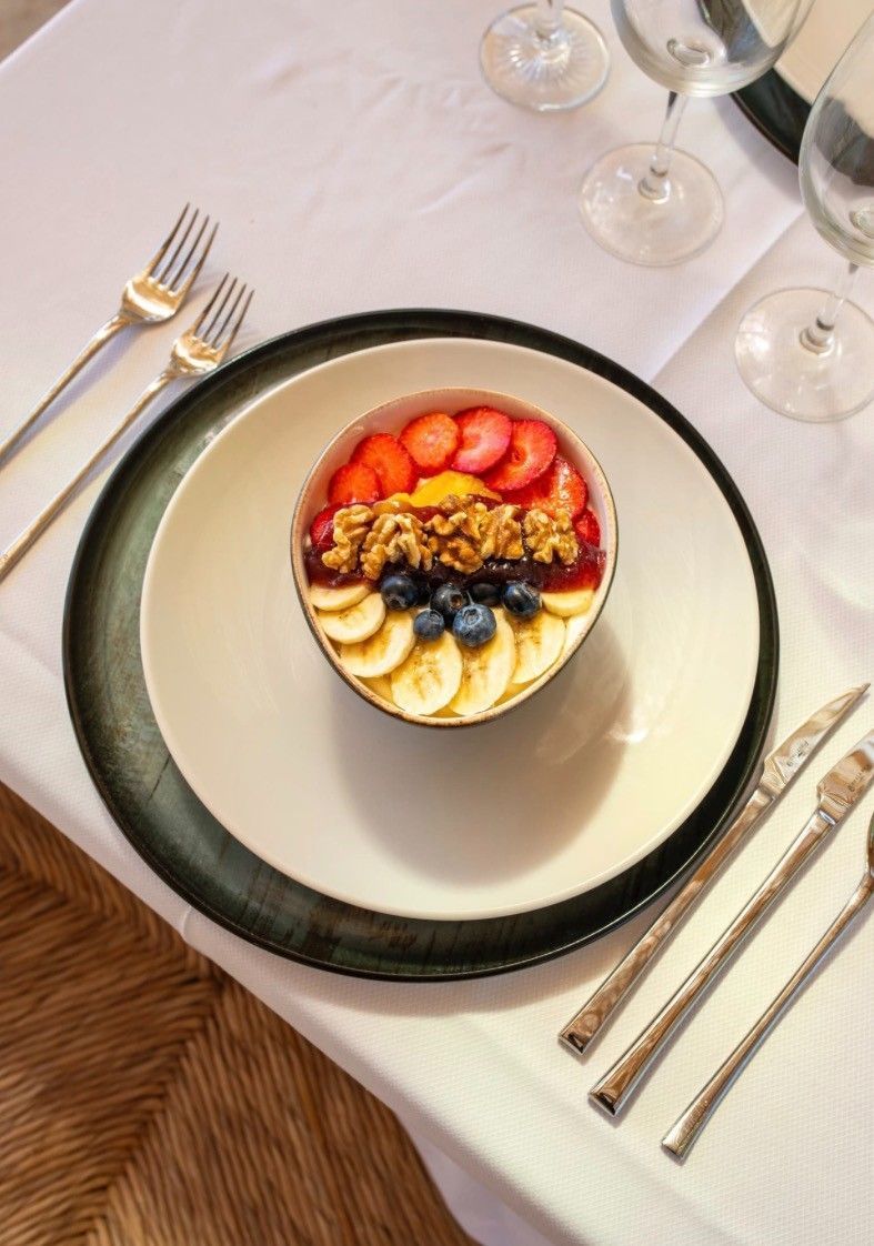 Postre con frutas en un cuenco blanco sobre una mesa puesta con cubiertos y copas de vino.