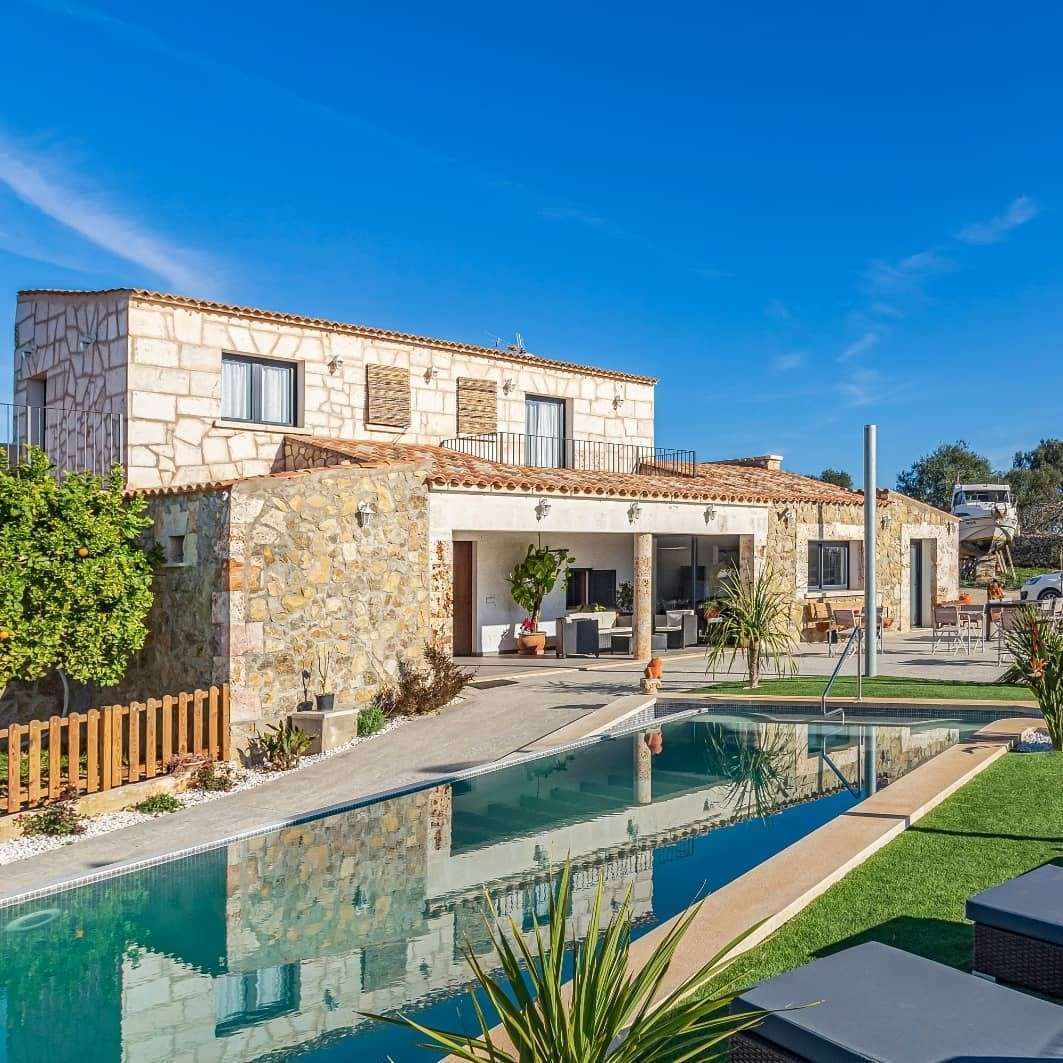 Villa moderna de piedra con piscina turquesa y patio bajo un cielo azul brillante.
