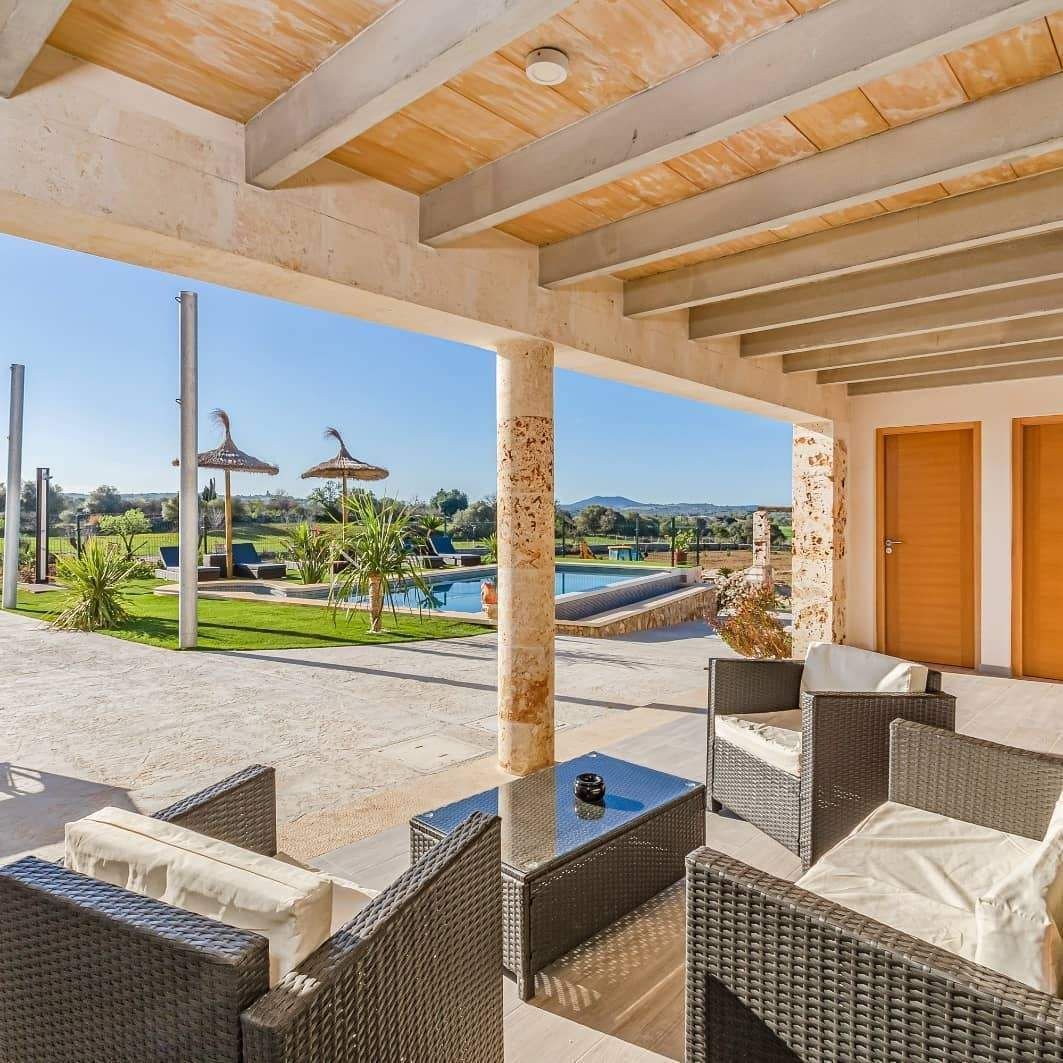 Patio cubierto con sillas de mimbre con vistas a una piscina soleada y a un jardín paisajístico del complejo.