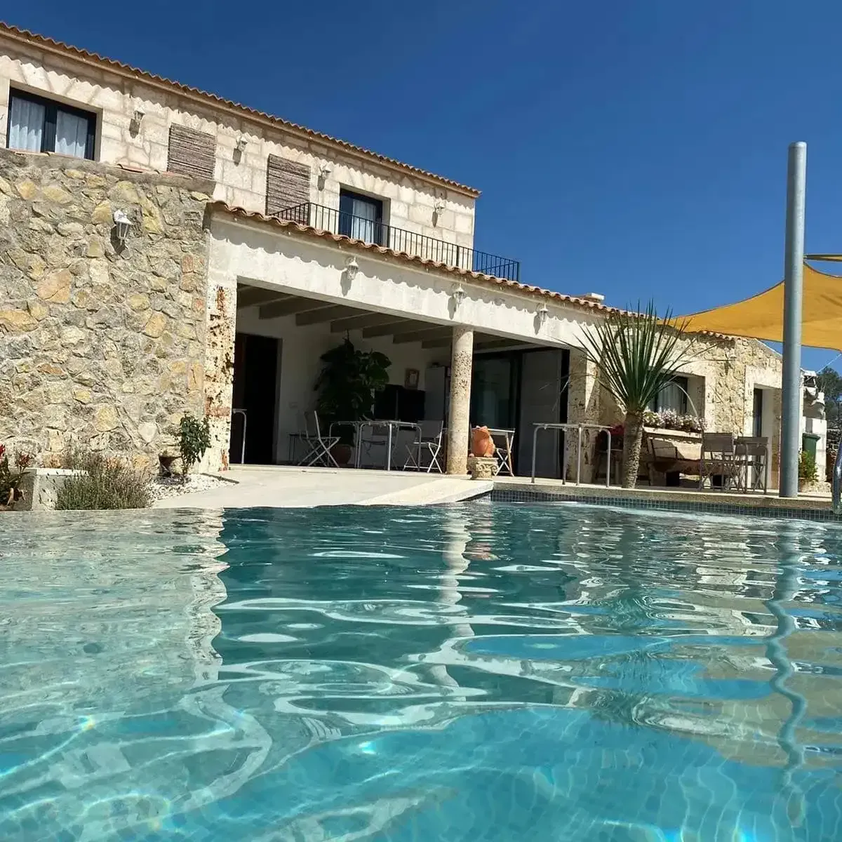 Villa de piedra con patio junto a la piscina y piscina azul bajo un cielo despejado.