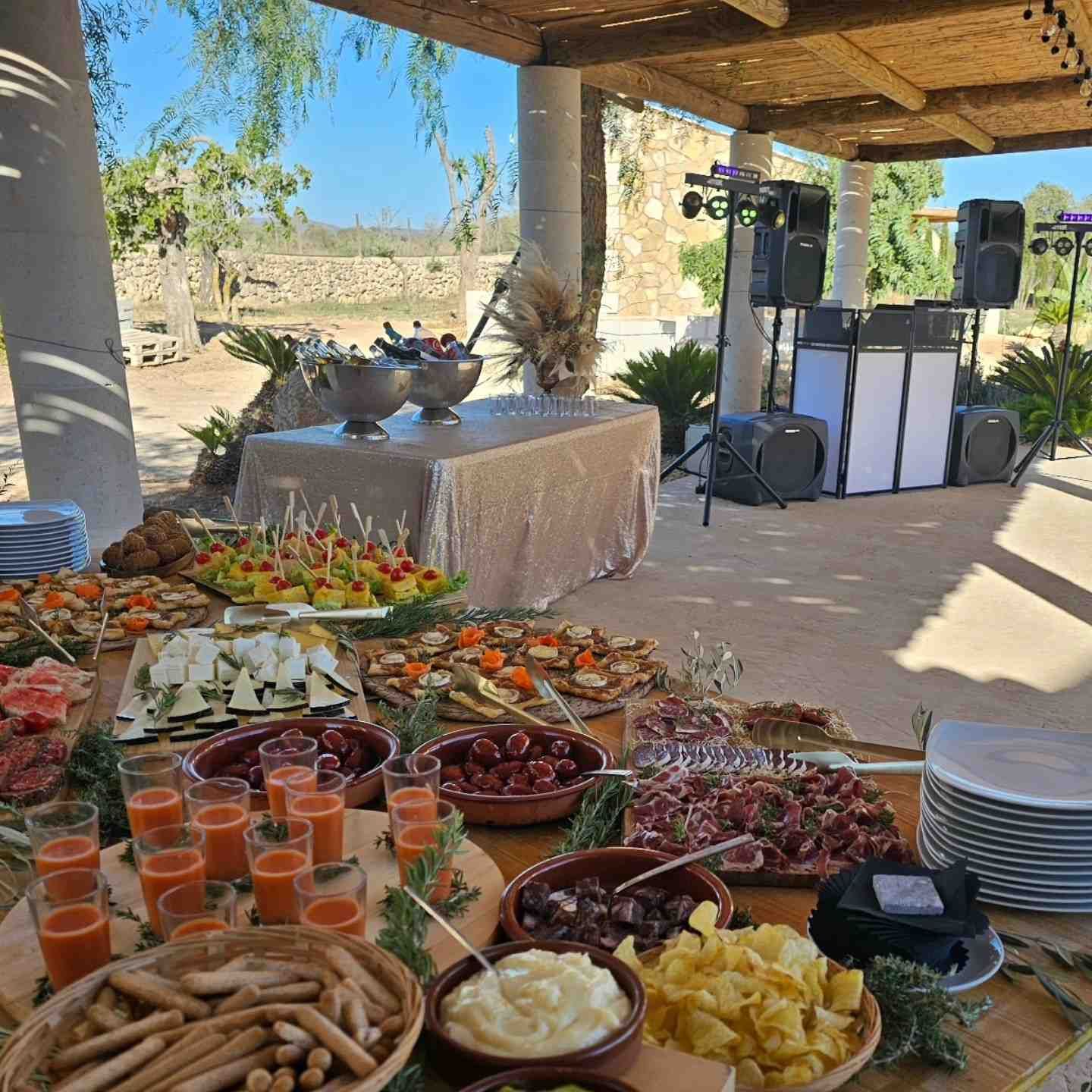 Mesa de buffet al aire libre con fruta, salsas y bebidas junto a altavoces bajo un patio sombreado con vista panorámica.