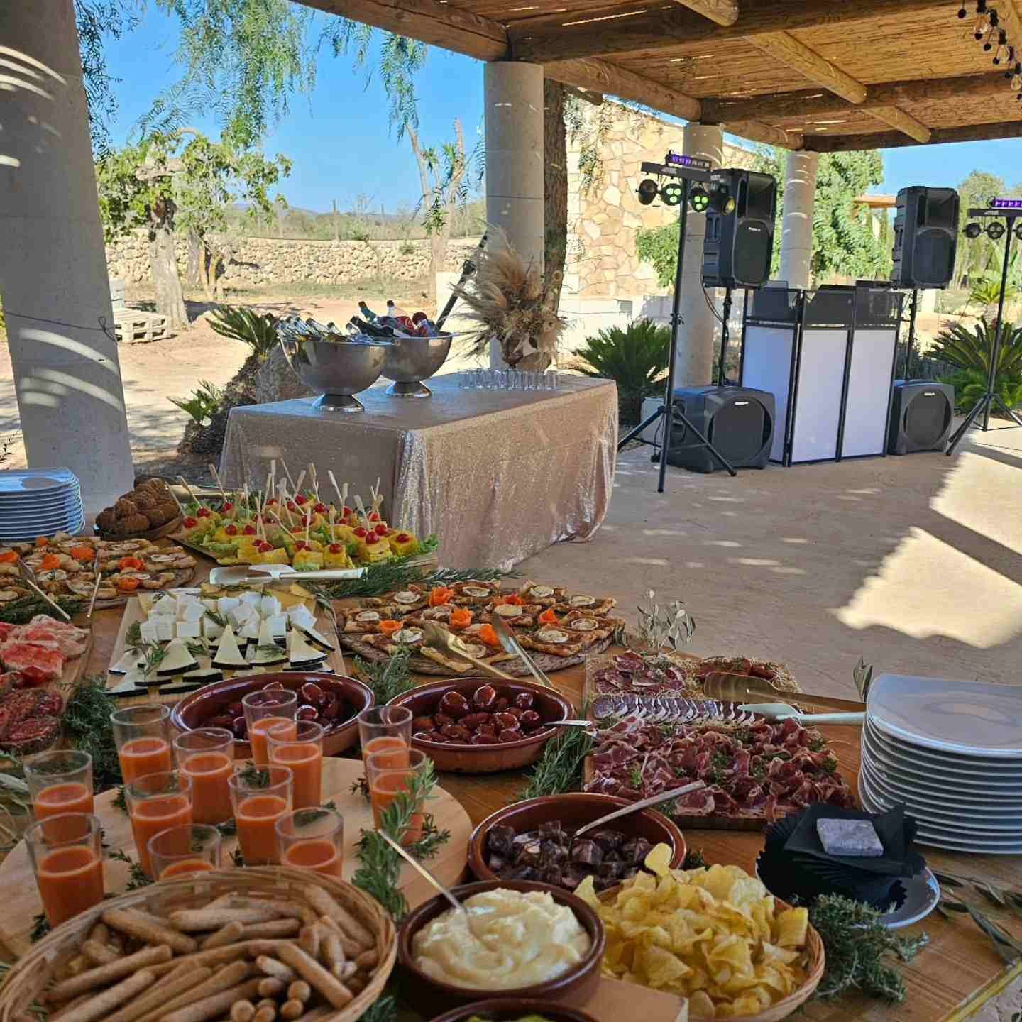 Mesa de buffet al aire libre con ensaladas, palitos de pan y bebidas bajo un patio sombreado junto a un sistema de sonido.