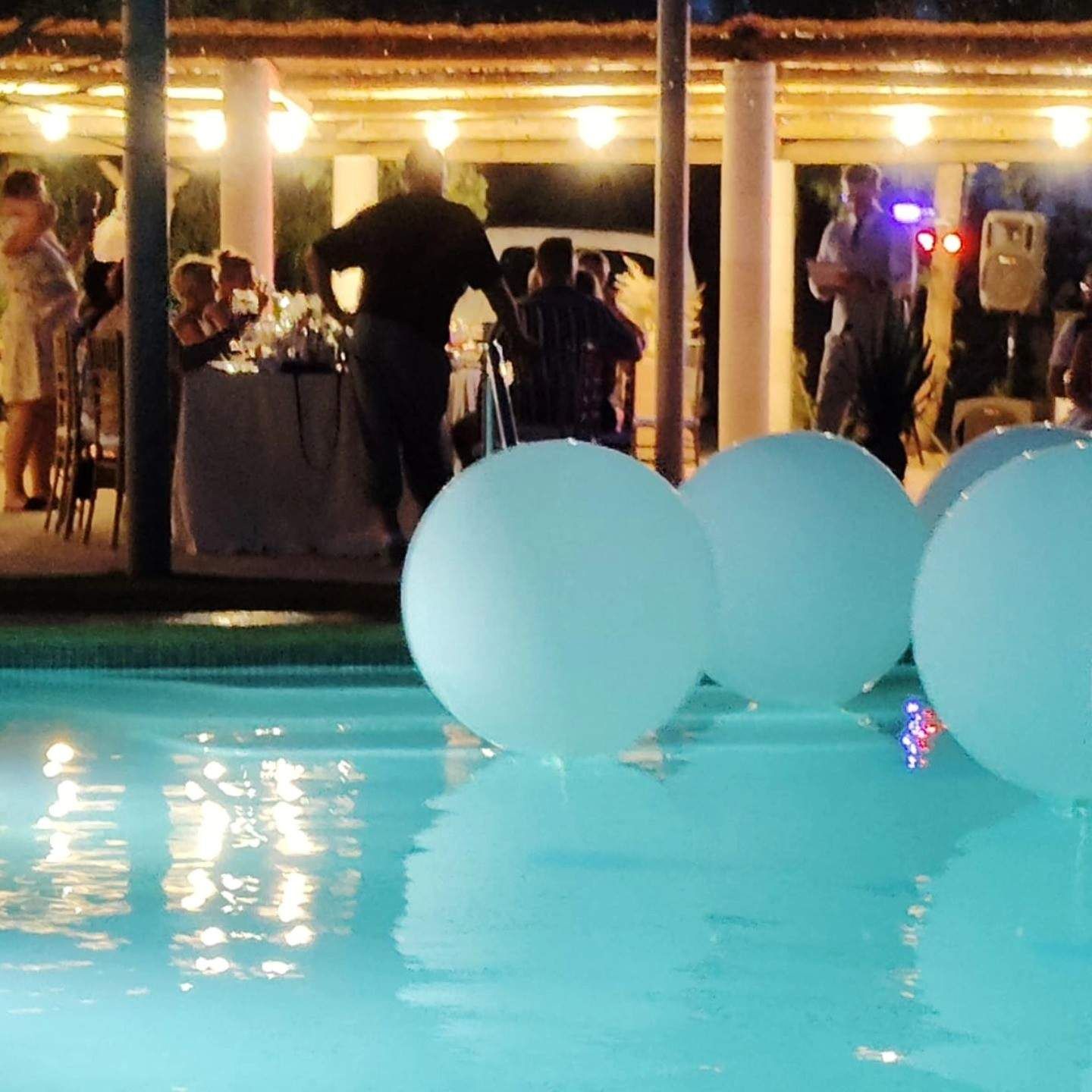 Evento nocturno junto a la piscina con grandes globos azul claro e invitados bajo luces cálidas.