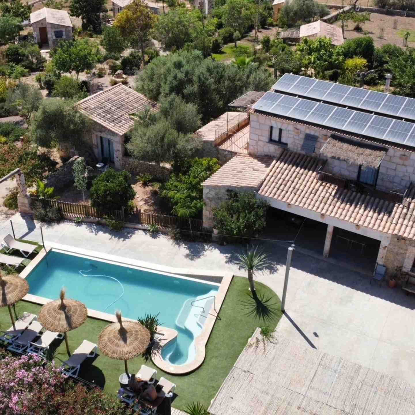 Vista aérea de una villa con piscina, sombrillas de patio y paneles solares rodeada de árboles.