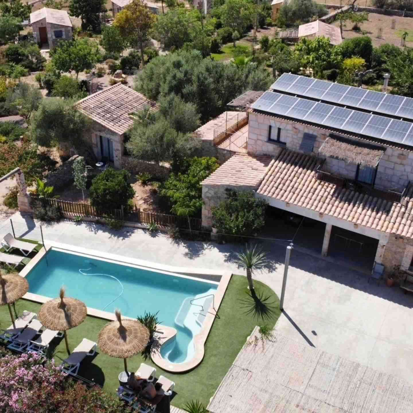 Vista aérea de una villa rústica con piscina, sombrillas de paja y paneles solares rodeada de vegetación.