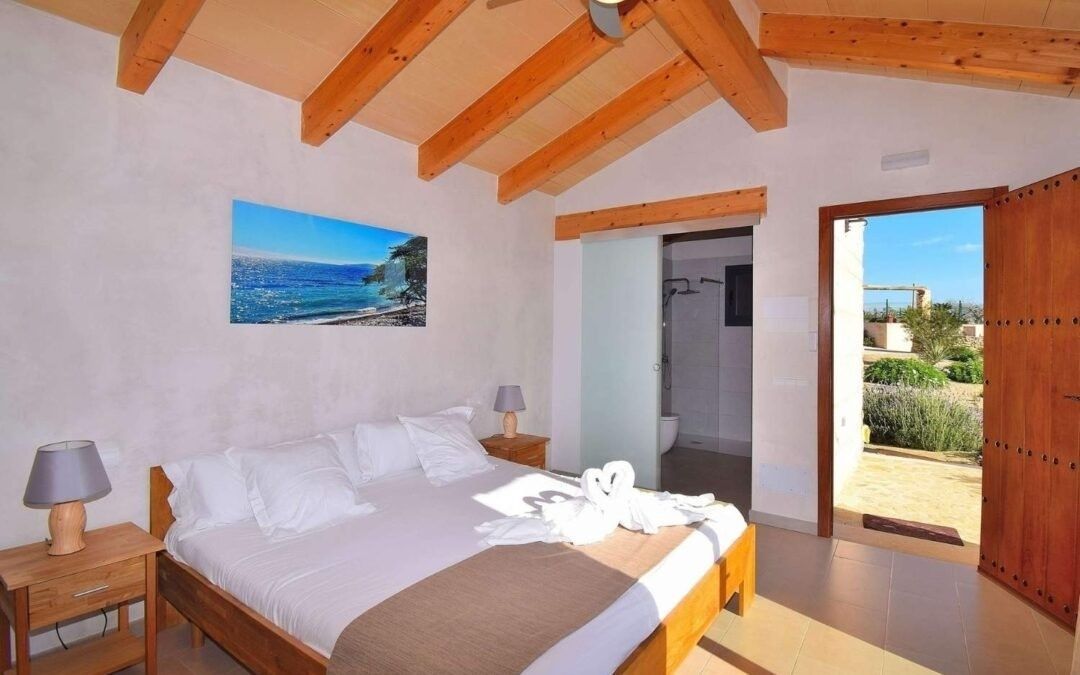 Habitación luminosa con vigas de madera en el techo, ropa de cama blanca y una puerta que da a un balcón soleado.