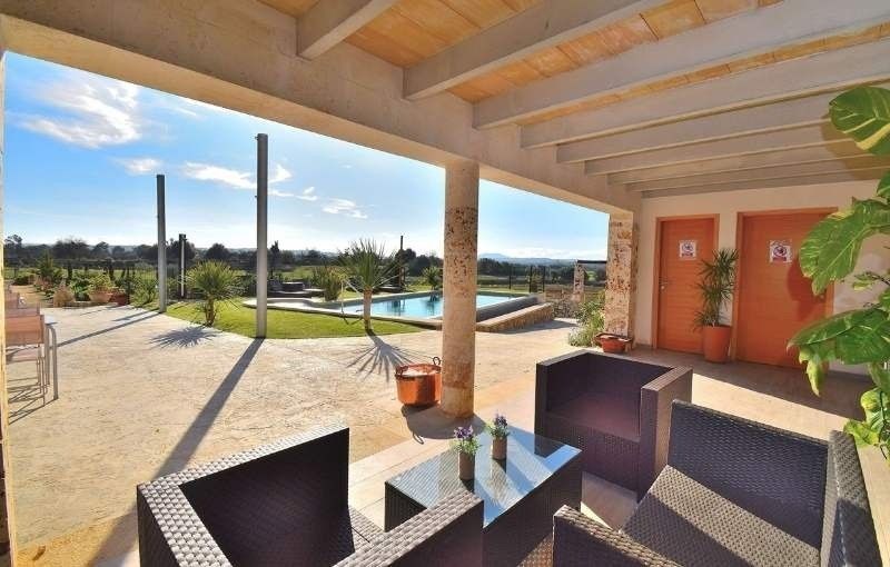Patio cubierto con asientos de mimbre, mesa de cristal, piscina y vistas al jardín soleado.