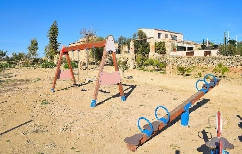 Un parque infantil vacío con columpios y un balancín en un patio polvoriento cerca de una casa.