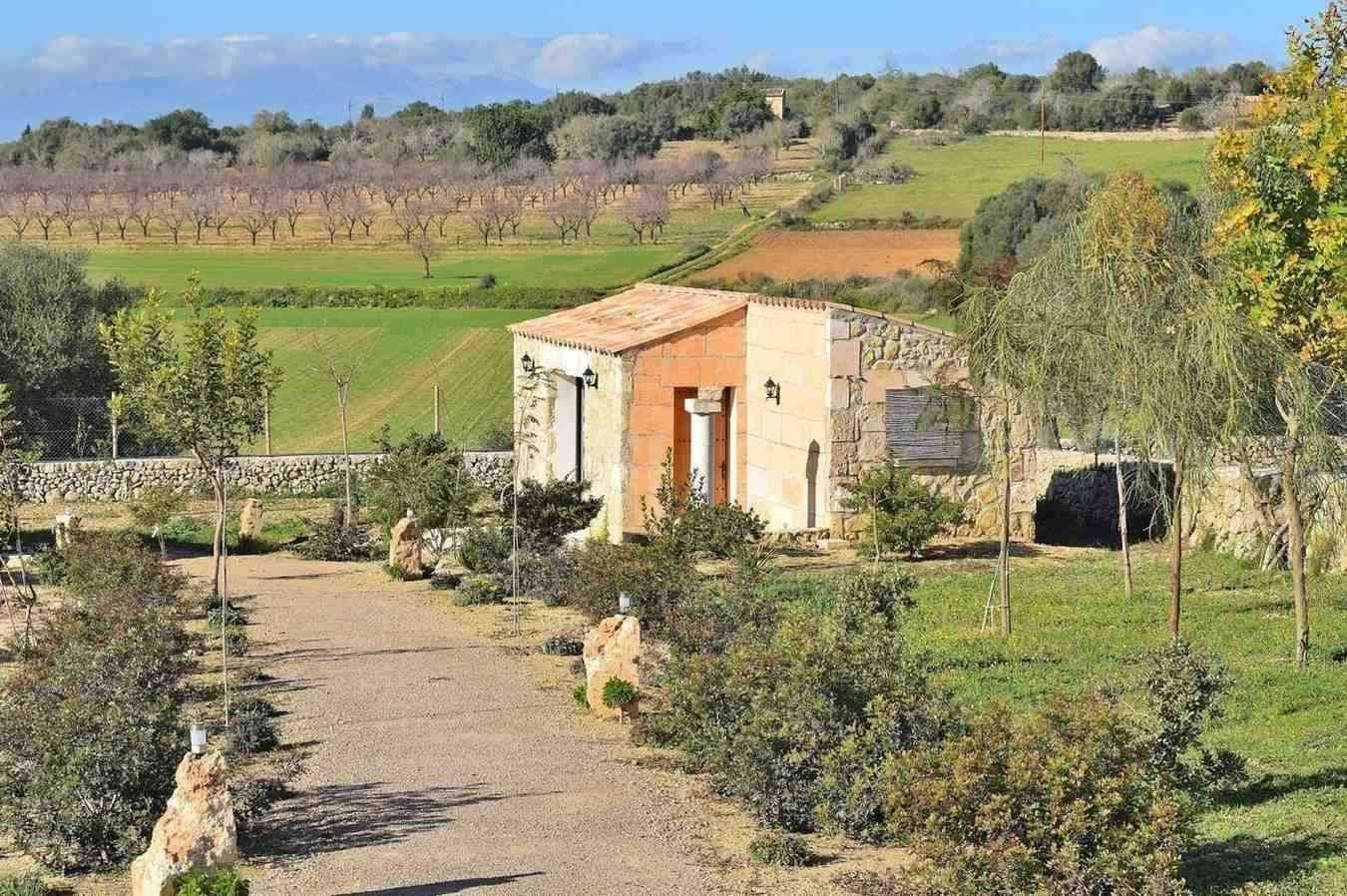Pequeña casa de piedra en un campo rural con árboles, arbustos y onduladas tierras de cultivo bajo un cielo azul.