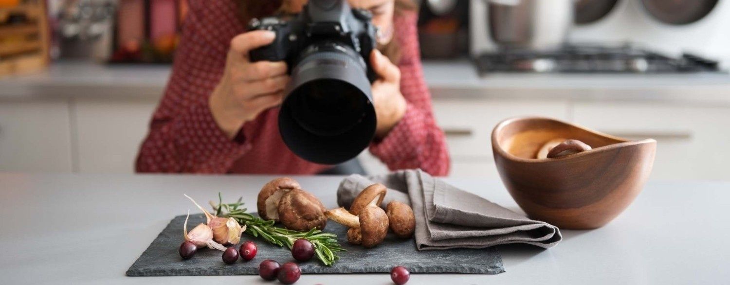 Fotógrafo encuadrando una naturaleza muerta de comida con pollo asado, uvas y cuencos sobre una mesa.