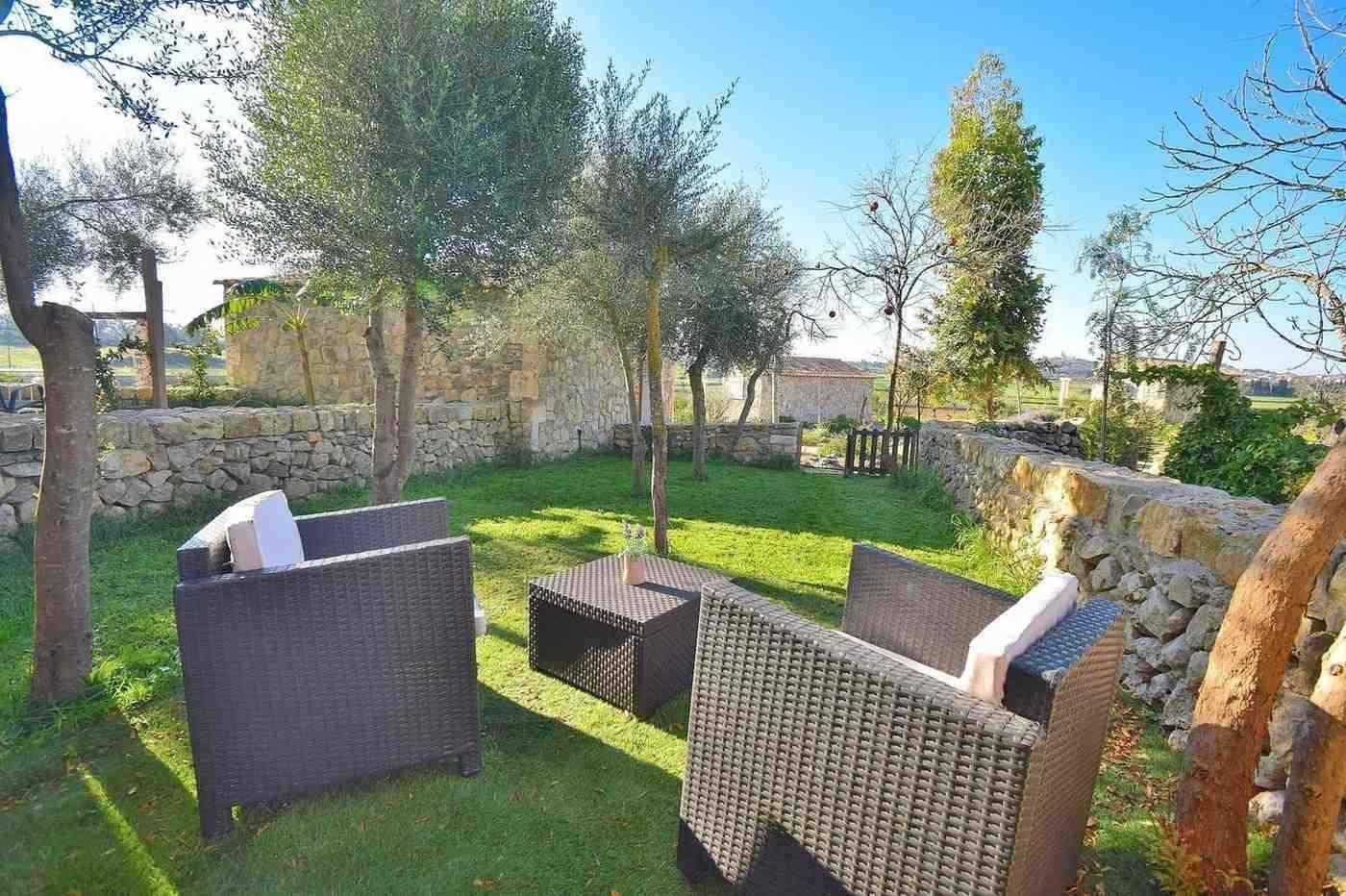 Patio soleado con sillas de mimbre, muros de piedra y árboles que rodean una pequeña mesa.