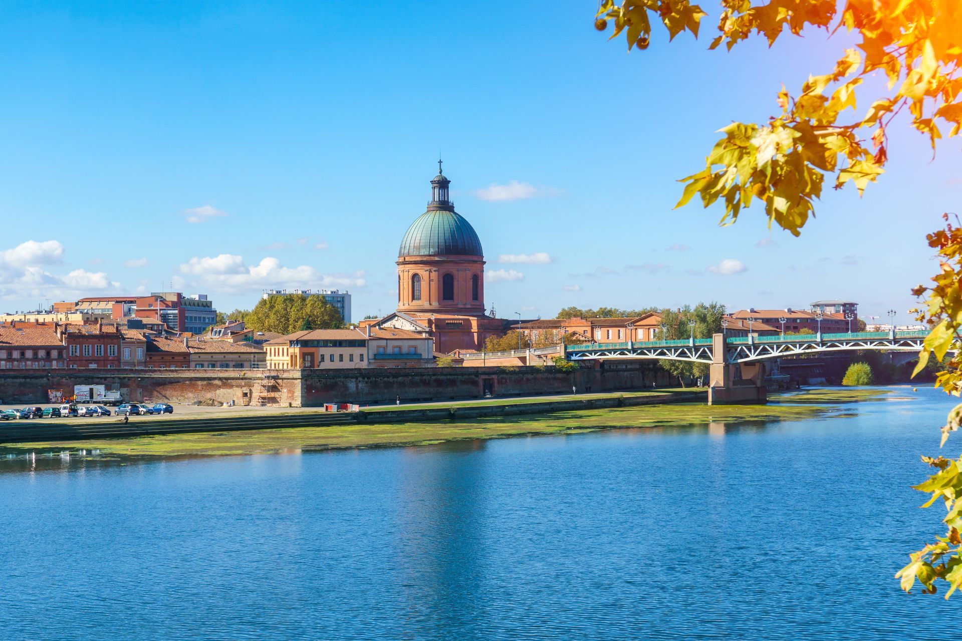 Paysage urbain de Toulouse avec des bâtiments en briques, une rivière bleue, un pont et une grande église en forme de dôme sous un ciel ensoleillé.