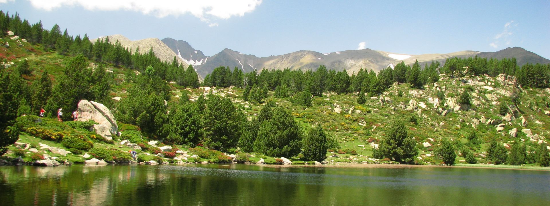 Un lac reflétant les arbres verts et la colline rocheuse sous un ciel bleu avec des montagnes enneigées.