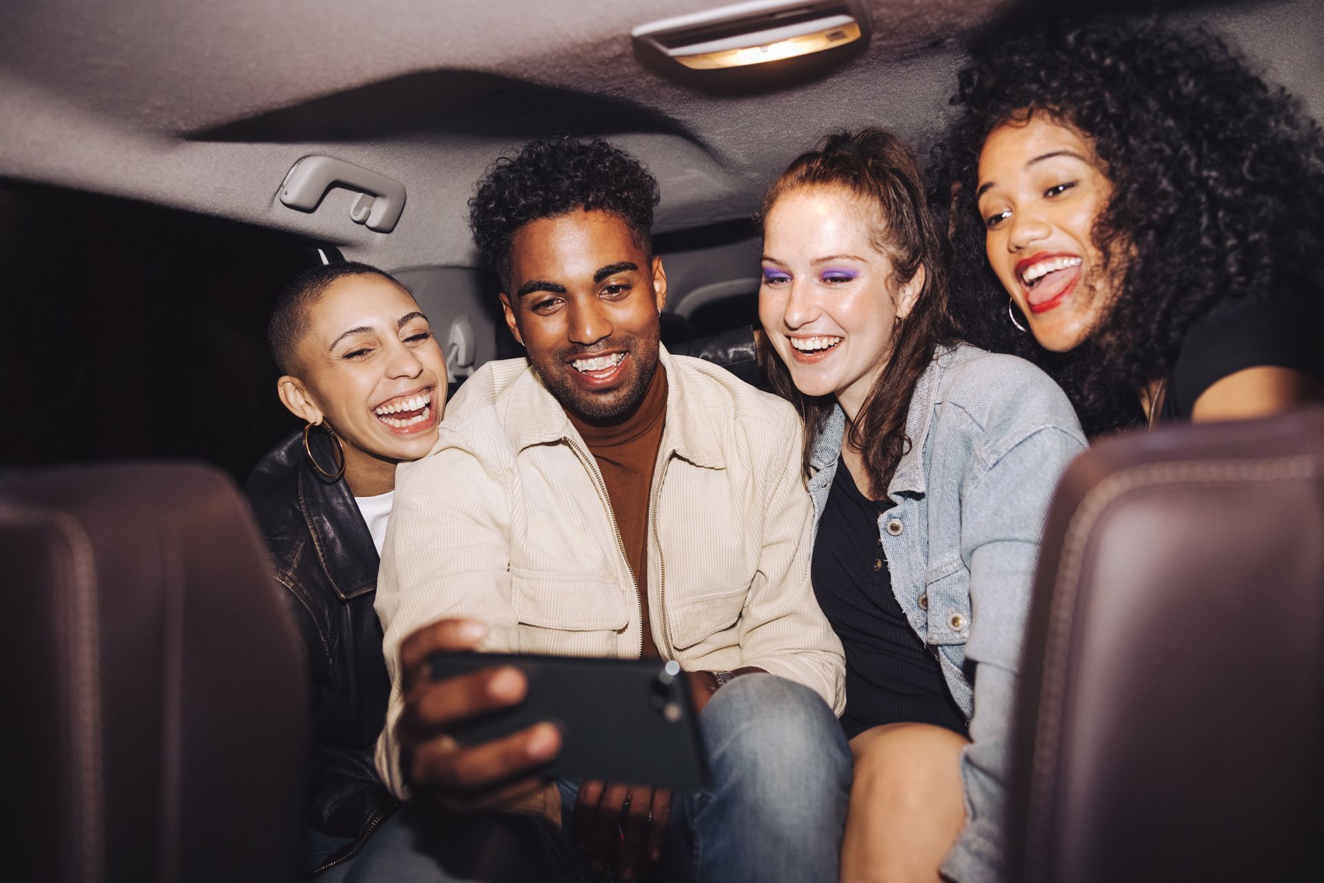 Quatre amis différents rient ensemble, regardant un téléphone à l'arrière d'une voiture la nuit.