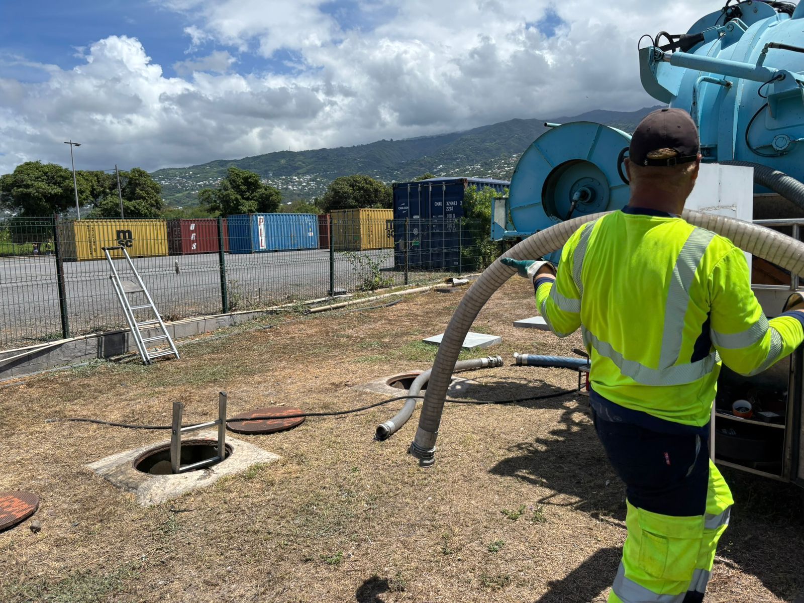 Professionnel en tenue jaune à La Réunion qui porte un tuyau vers une bouche d'égout ouverte.