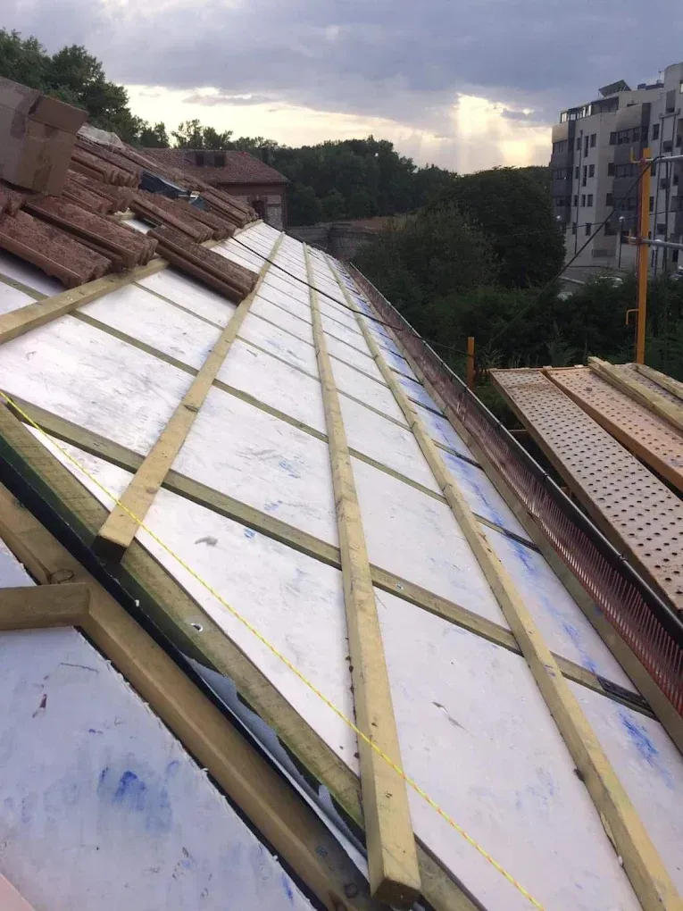Techo en construcción con paneles aislantes blancos, listones de madera y pilas de tejas marrones contra el cielo de fondo.