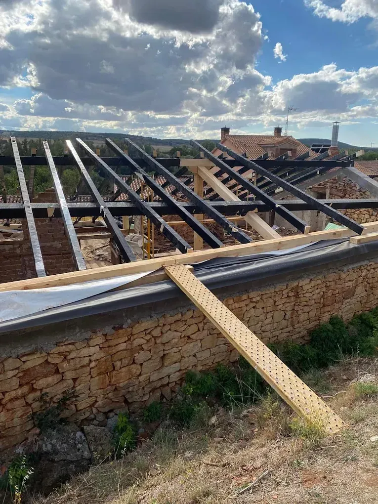 Obra en construcción con vigas de acero negro a la vista, refuerzos de madera y un muro de piedra bajo un cielo azul nublado.
